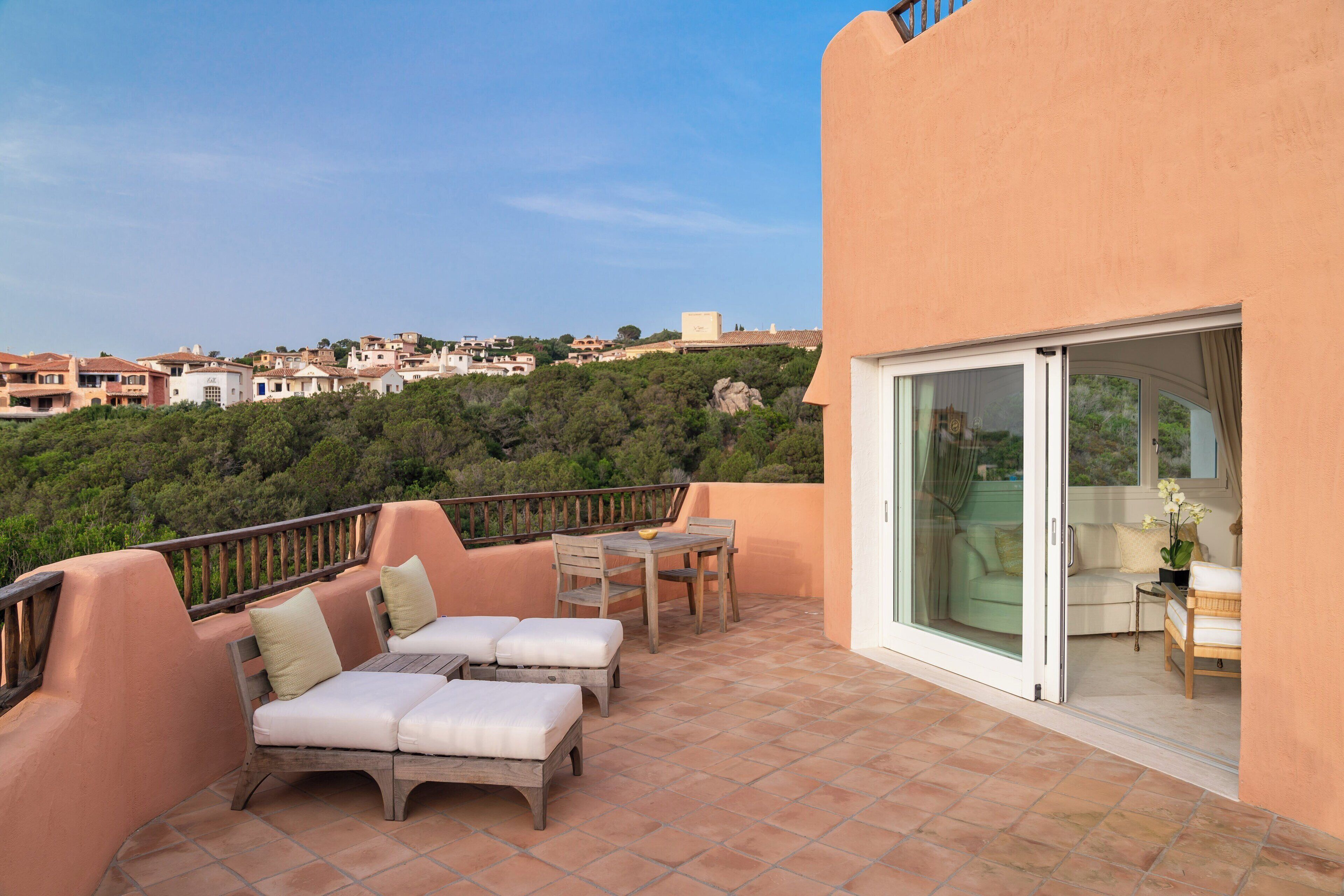 A terrace with outdoor furniture, a glass door leading to a room, and a view of a hillside with buildings.