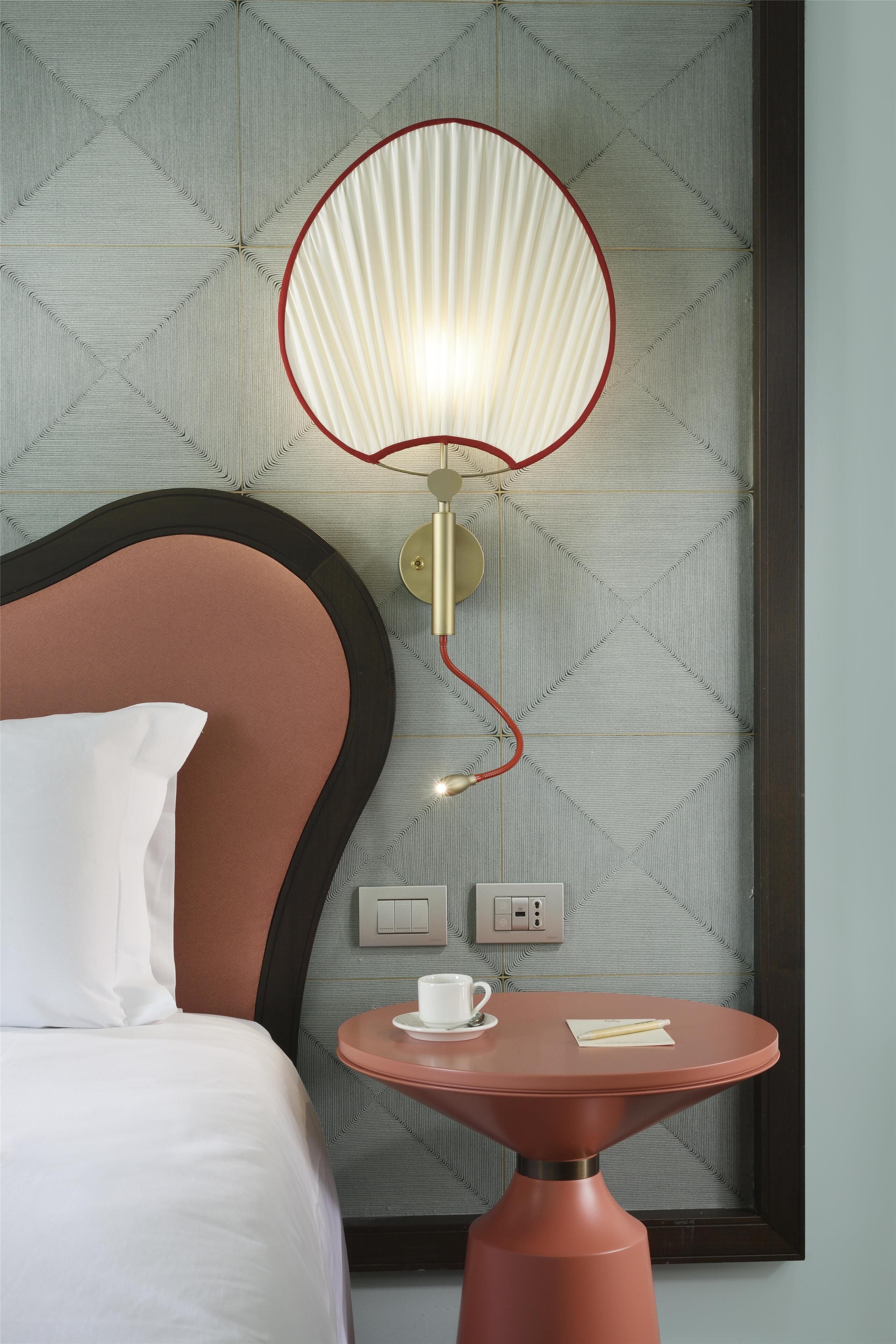 A hotel room with a bed, a table with a cup and a notepad, and a wall-mounted lamp.