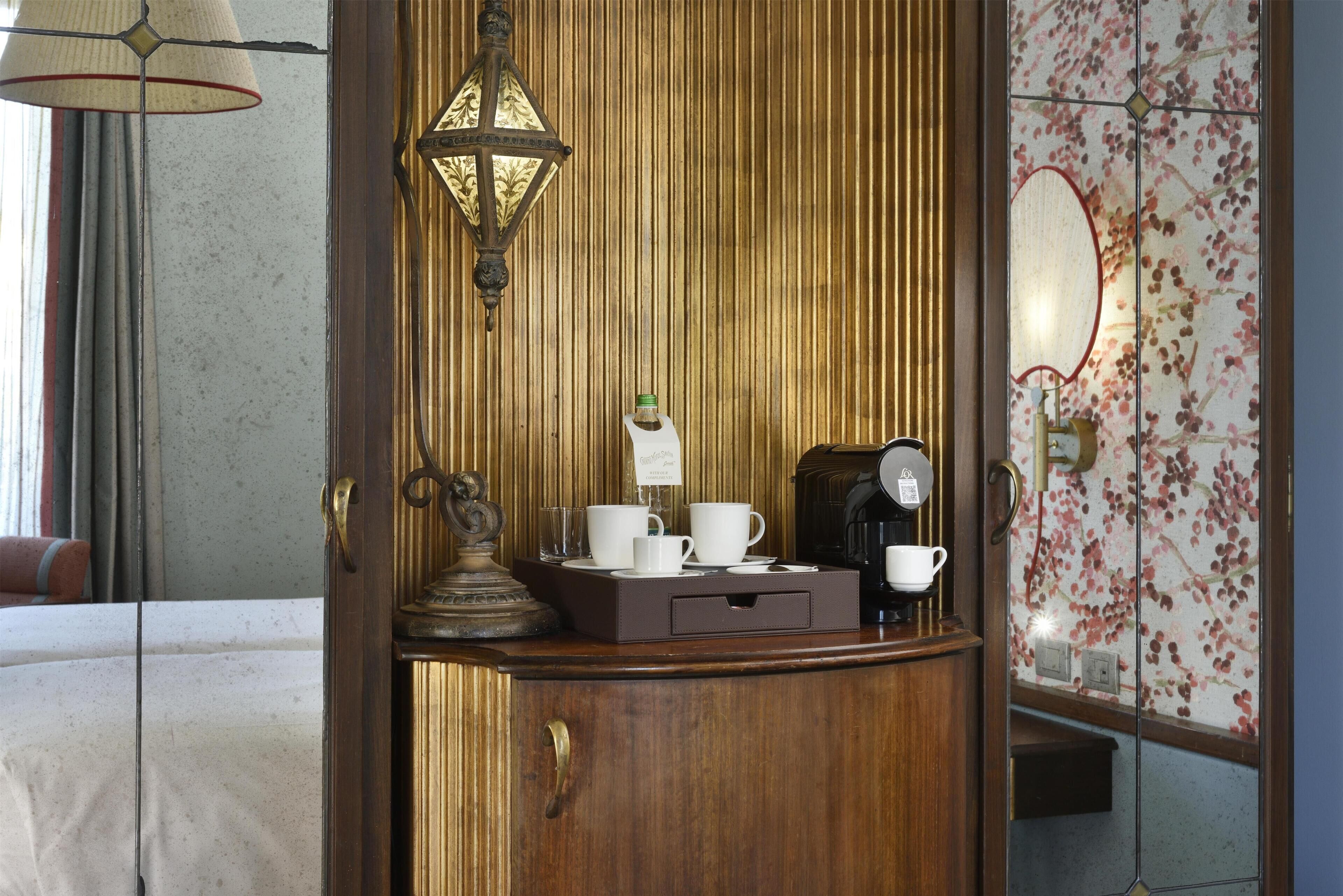 A wooden counter with a coffee maker, cups, and a tray, a mirror, and a lamp.