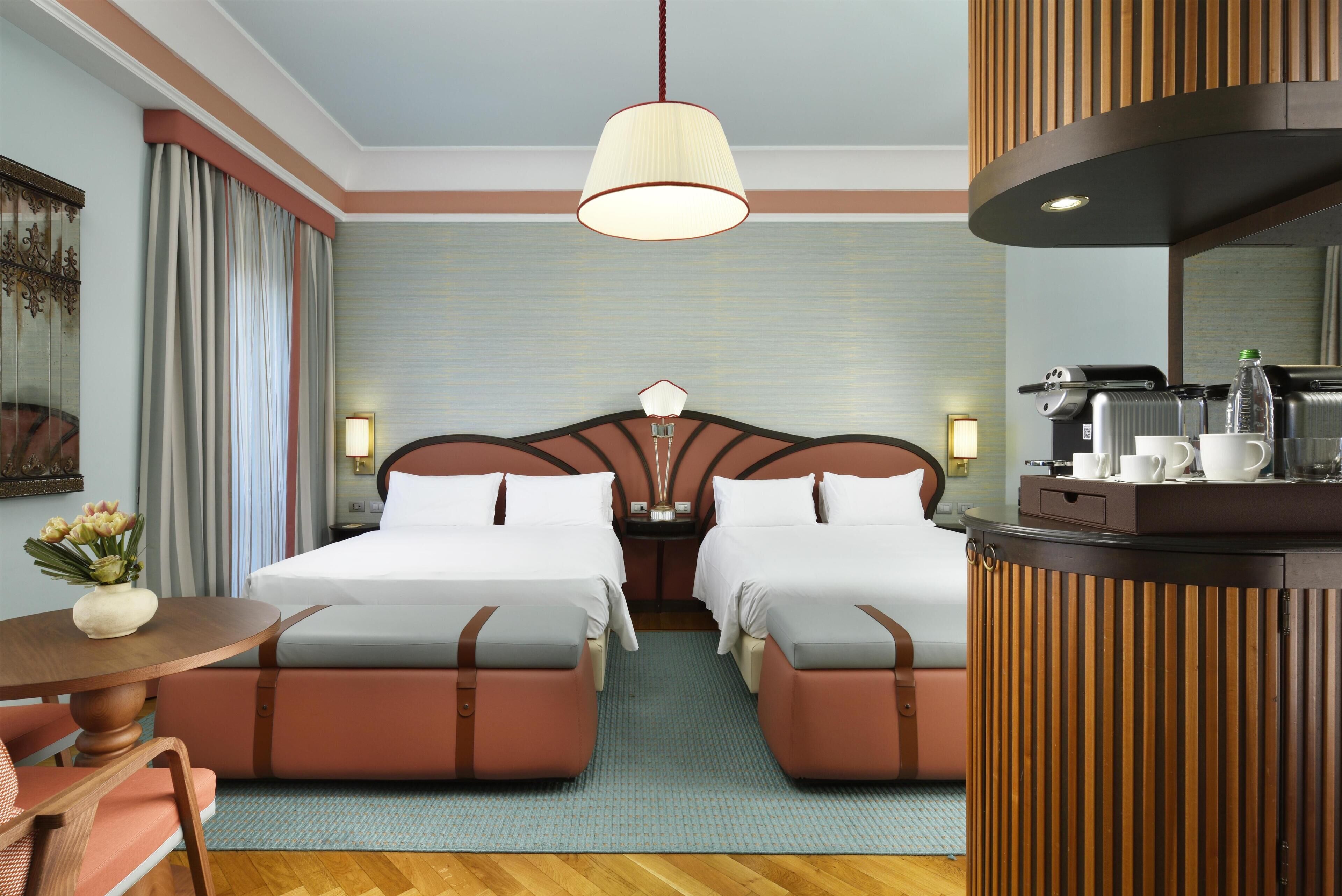 A hotel room with two beds, a desk with a chair, a coffee machine, and a mirror.