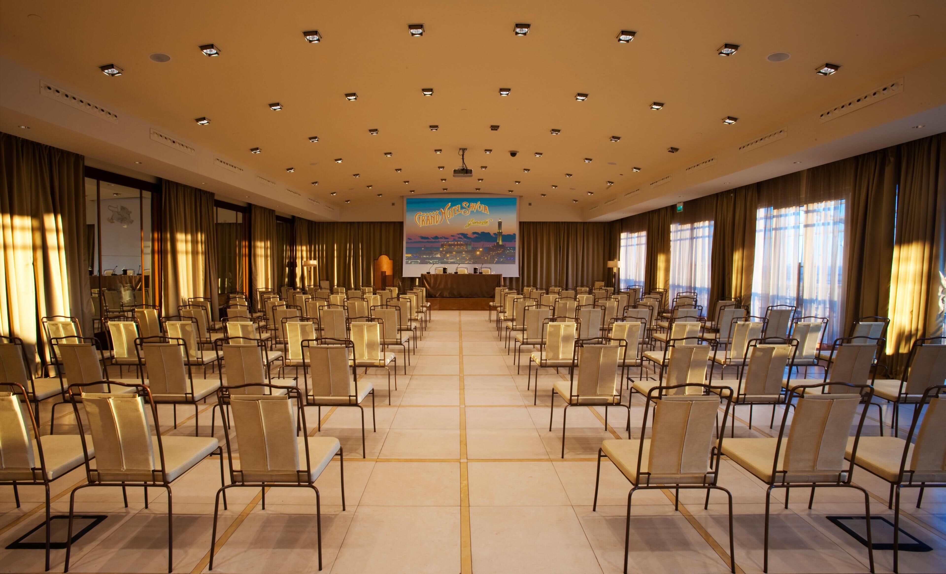 Une salle de conférence avec des rangées de chaises, une scène et un grand écran sur lequel est projetée une vue panoramique de la ville.