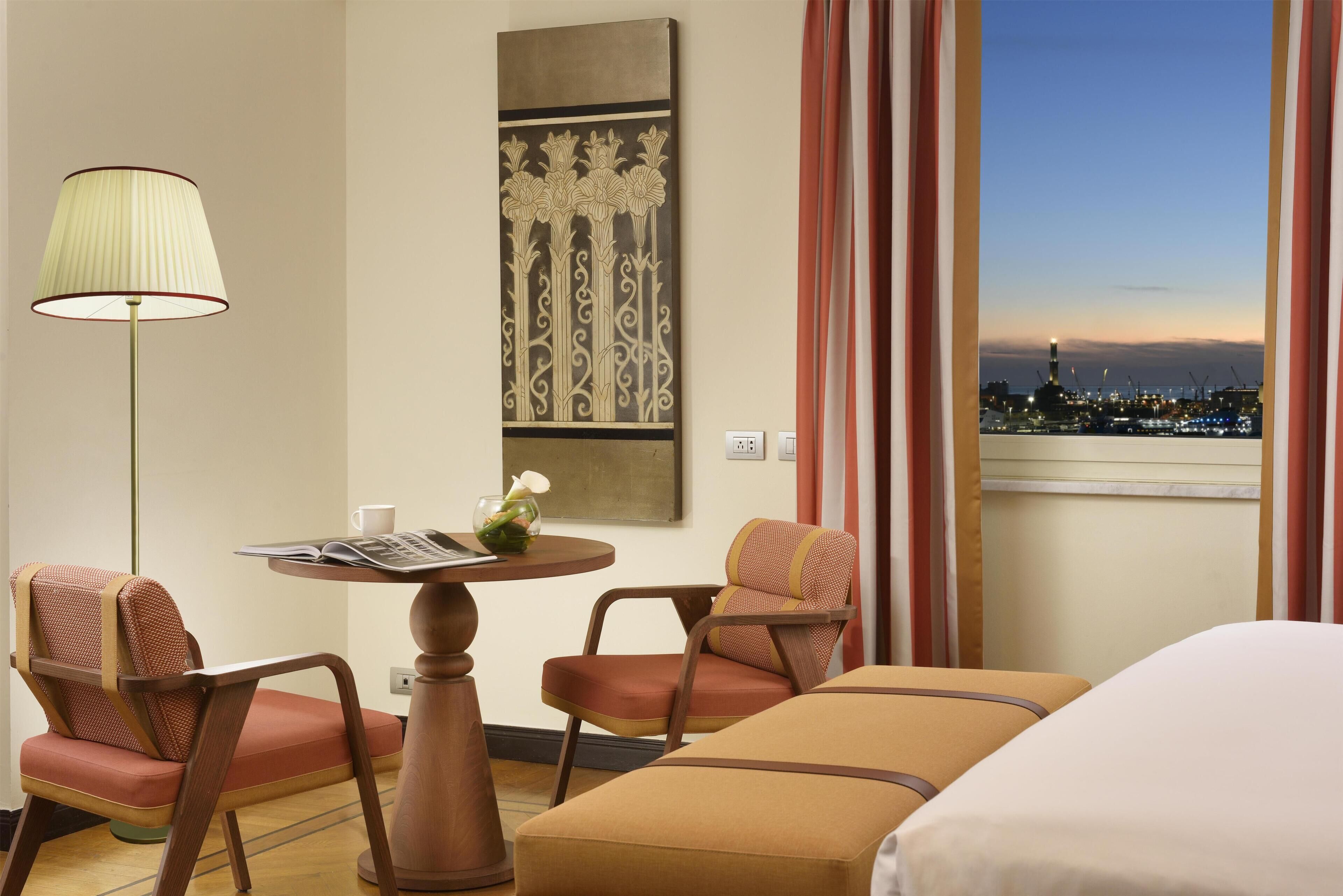 A hotel room with a bed, two armchairs, a table with a lamp, a vase with flowers, and a painting on the wall.