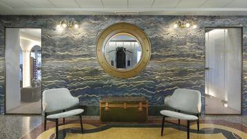 A lobby with a circular mirror, two armchairs, a vintage suitcase, and a patterned wall.
