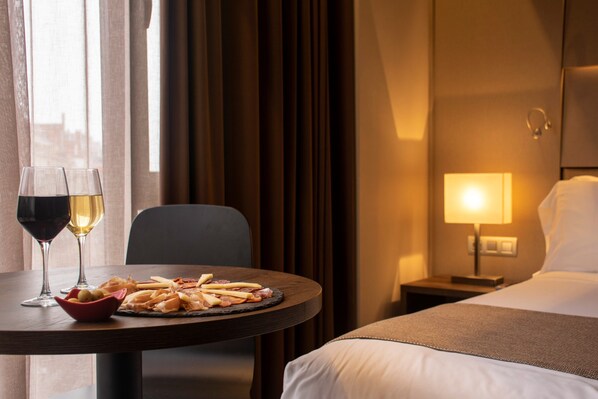 Une chambre d’hôtel avec un lit, une table de chevet avec une lampe, une table à manger avec une assiette de nourriture et des verres de vin.