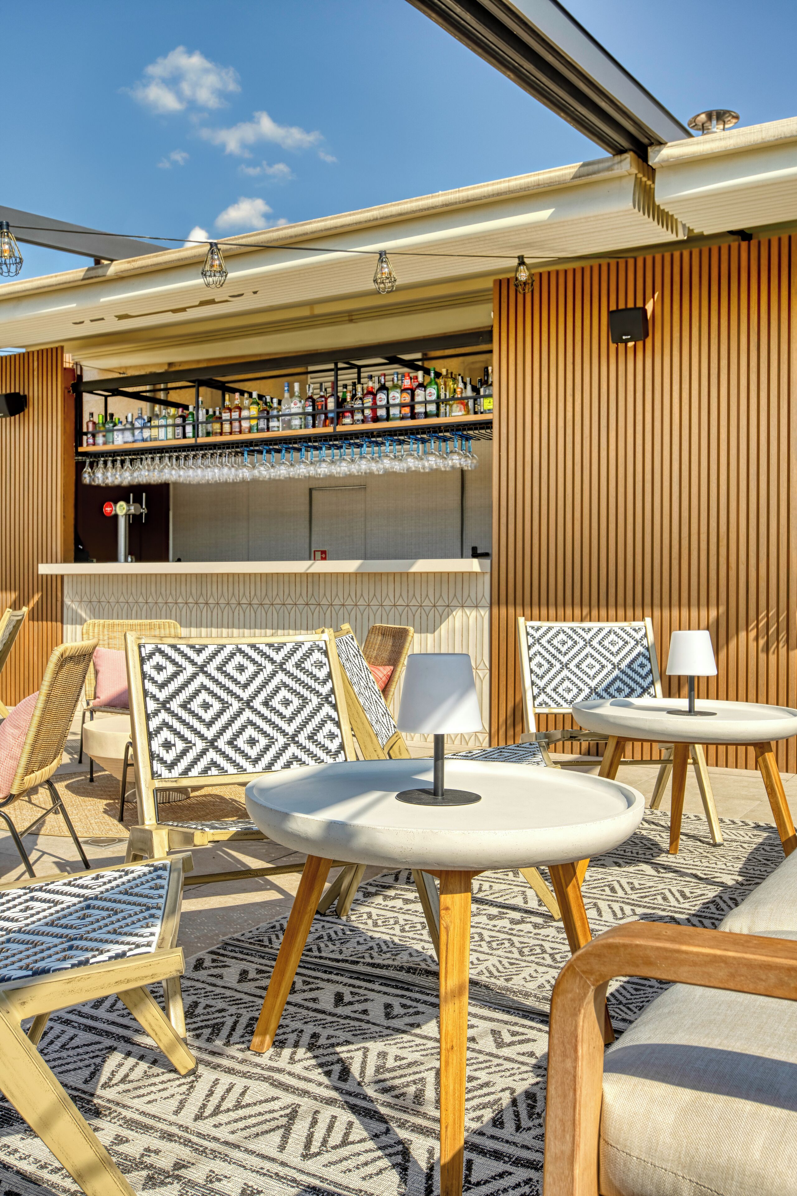 Een bar met houten lambrisering, een bargedeelte en diverse flessen en glazen. Er staan verschillende stoelen en tafels met decoratieve kussens.