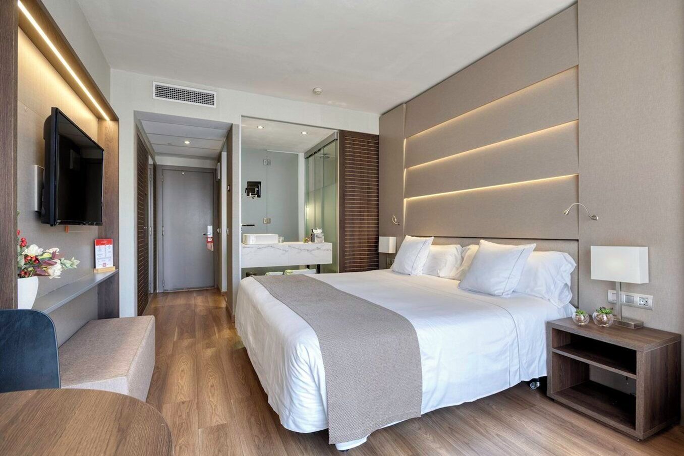 Una habitación de hotel moderna con una cama grande, mesitas de noche, televisión y baño con espejo y lavabo.