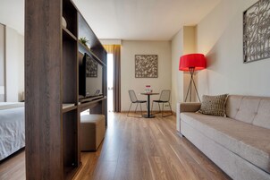 A modern hotel room with a wooden floor, a sofa, a TV, a small table with chairs, and artwork on the walls.