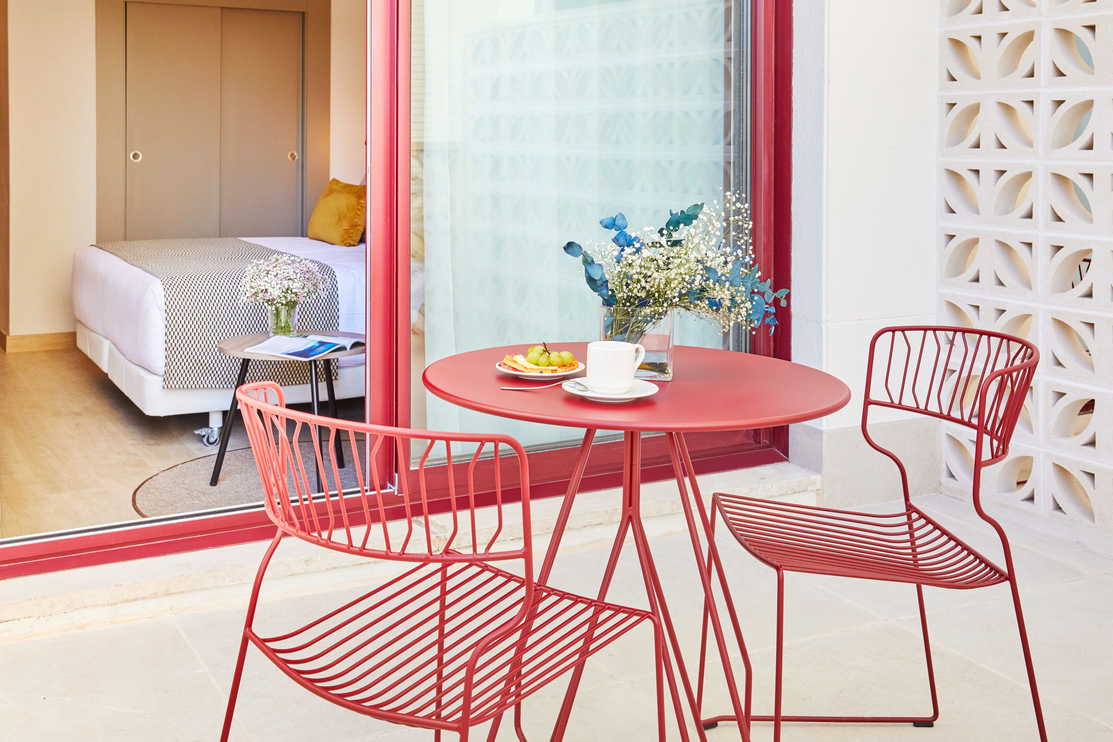 A modern hotel room with a bed, a small table with a vase of flowers, and red metal chairs.