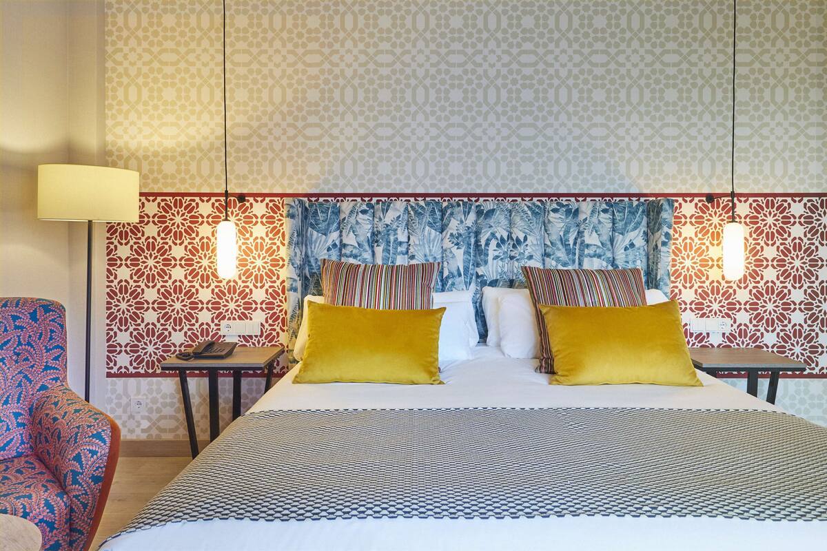 A hotel room with a patterned headboard, a bed with two pillows, a nightstand, a lamp, and a chair.