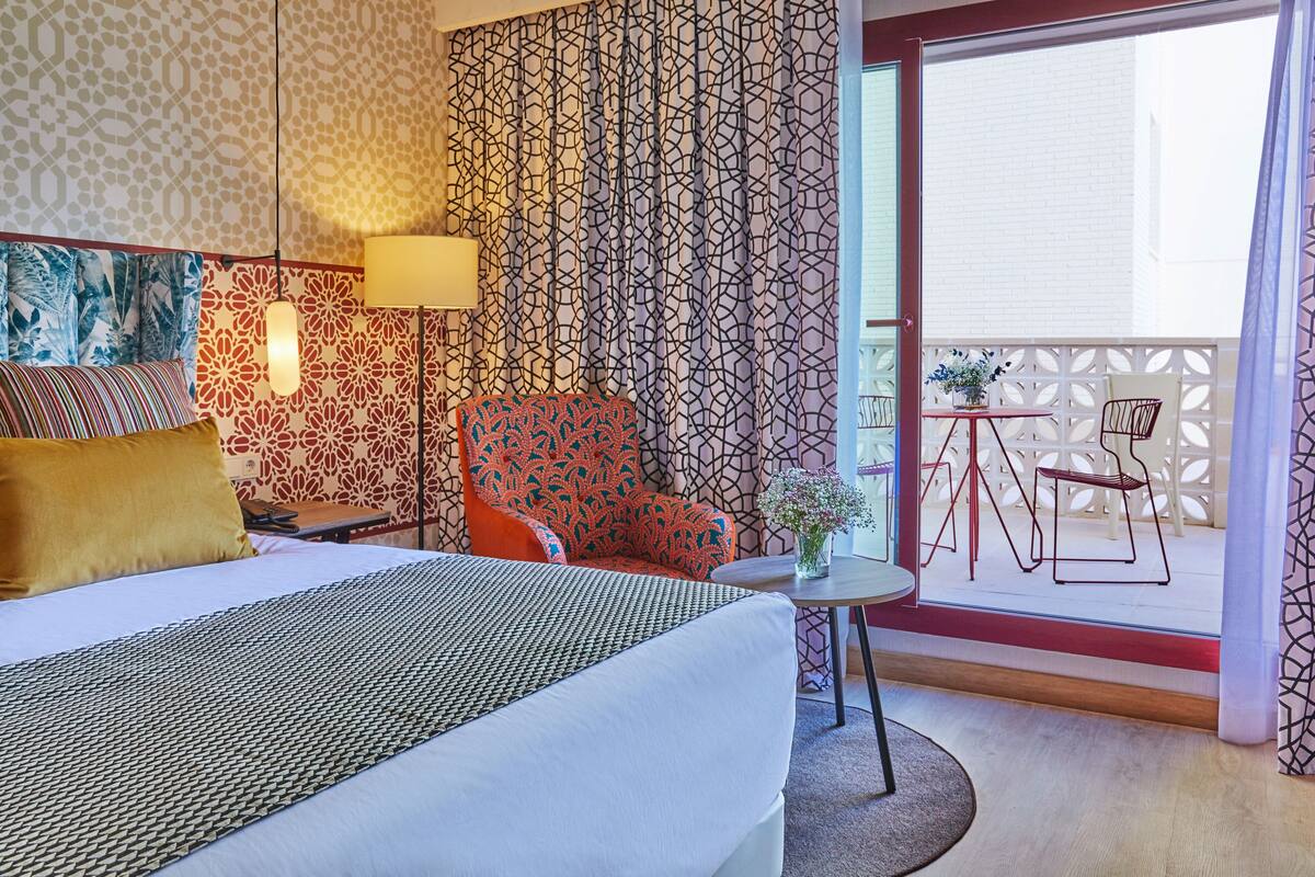A hotel room with a bed, a chair, a small table with a vase, and a balcony with outdoor furniture.