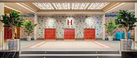 A hotel lobby with a patterned floral wall, a reception desk, and potted plants.