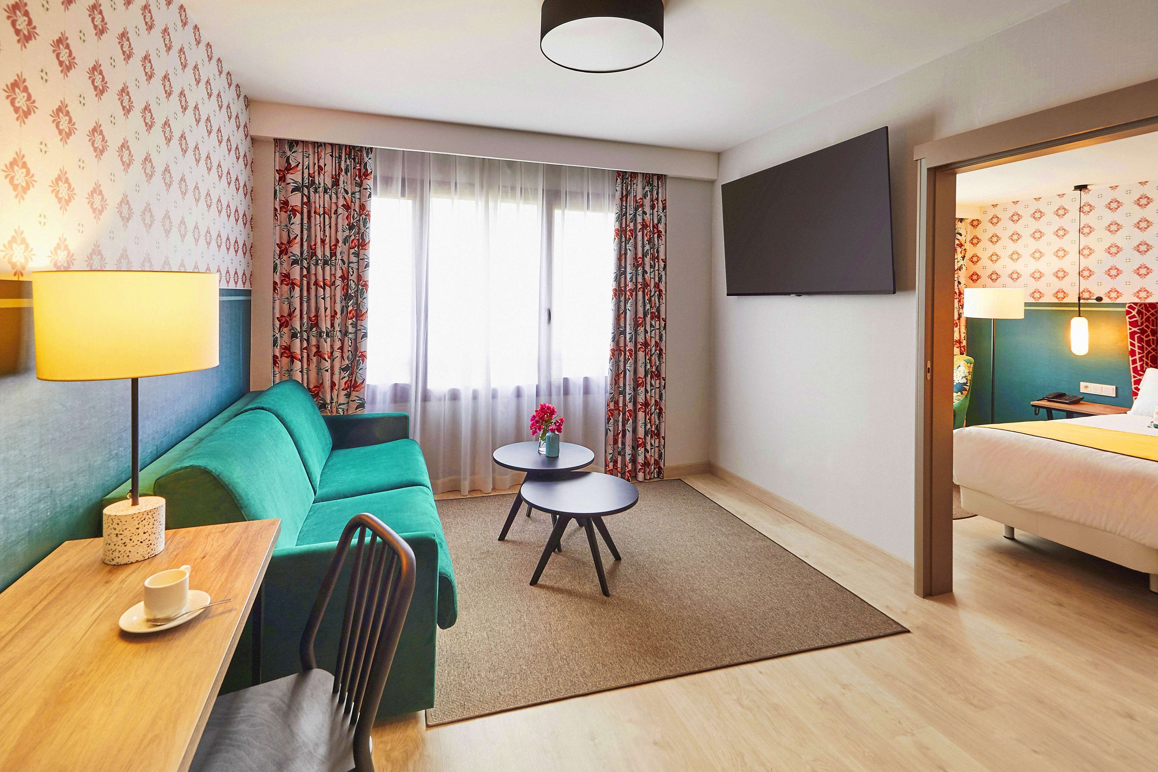 A hotel room with a green sofa, a small round table with a lamp, a bed, and a TV mounted on the wall.