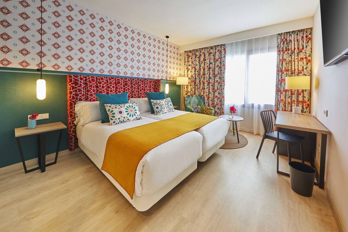 A hotel room with a bed, a desk, a chair, a small table, and a TV.