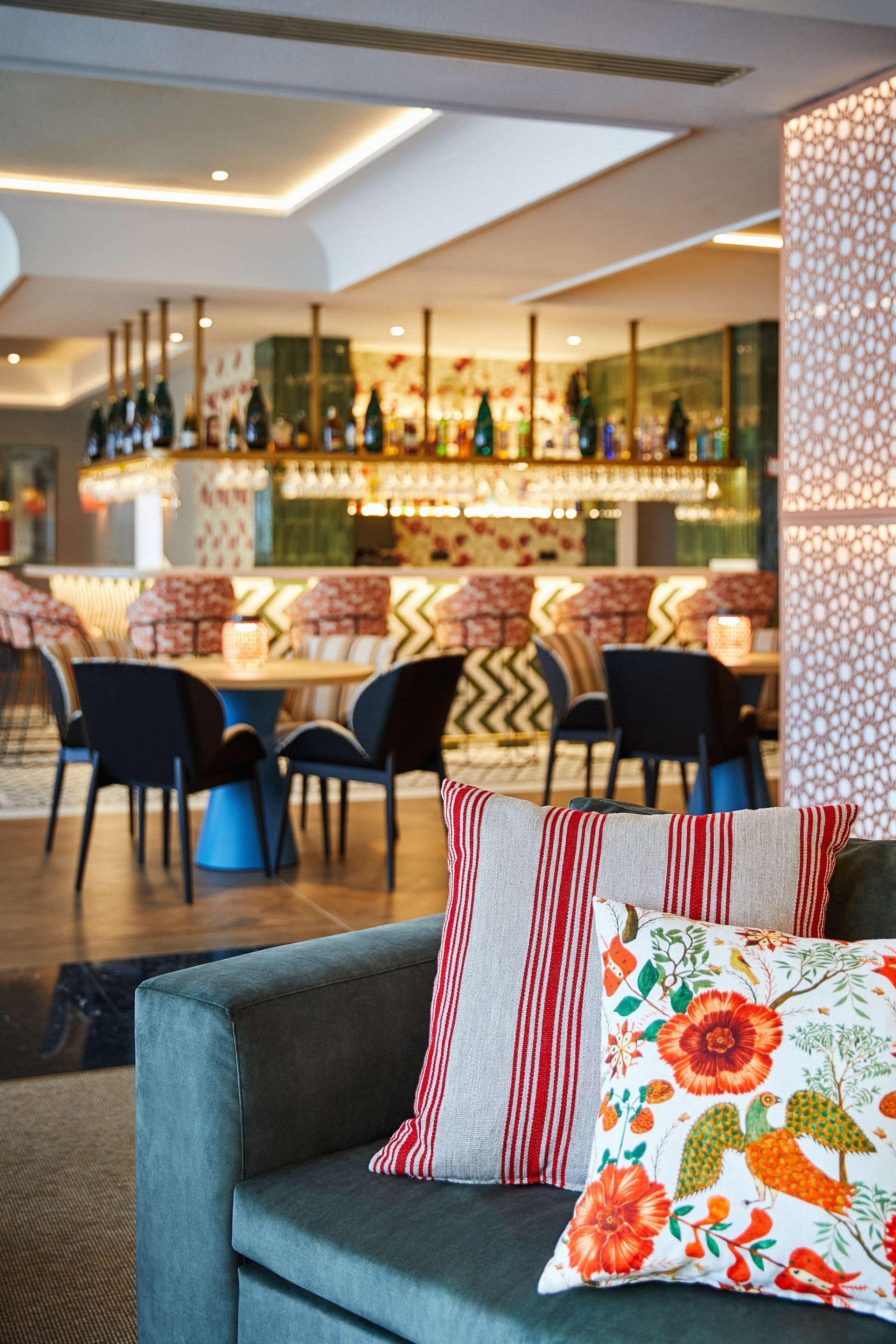 Un hall d’hôtel moderne avec un bar, des espaces aménagés pour s’asseoir et des coussins décoratifs sur un canapé.