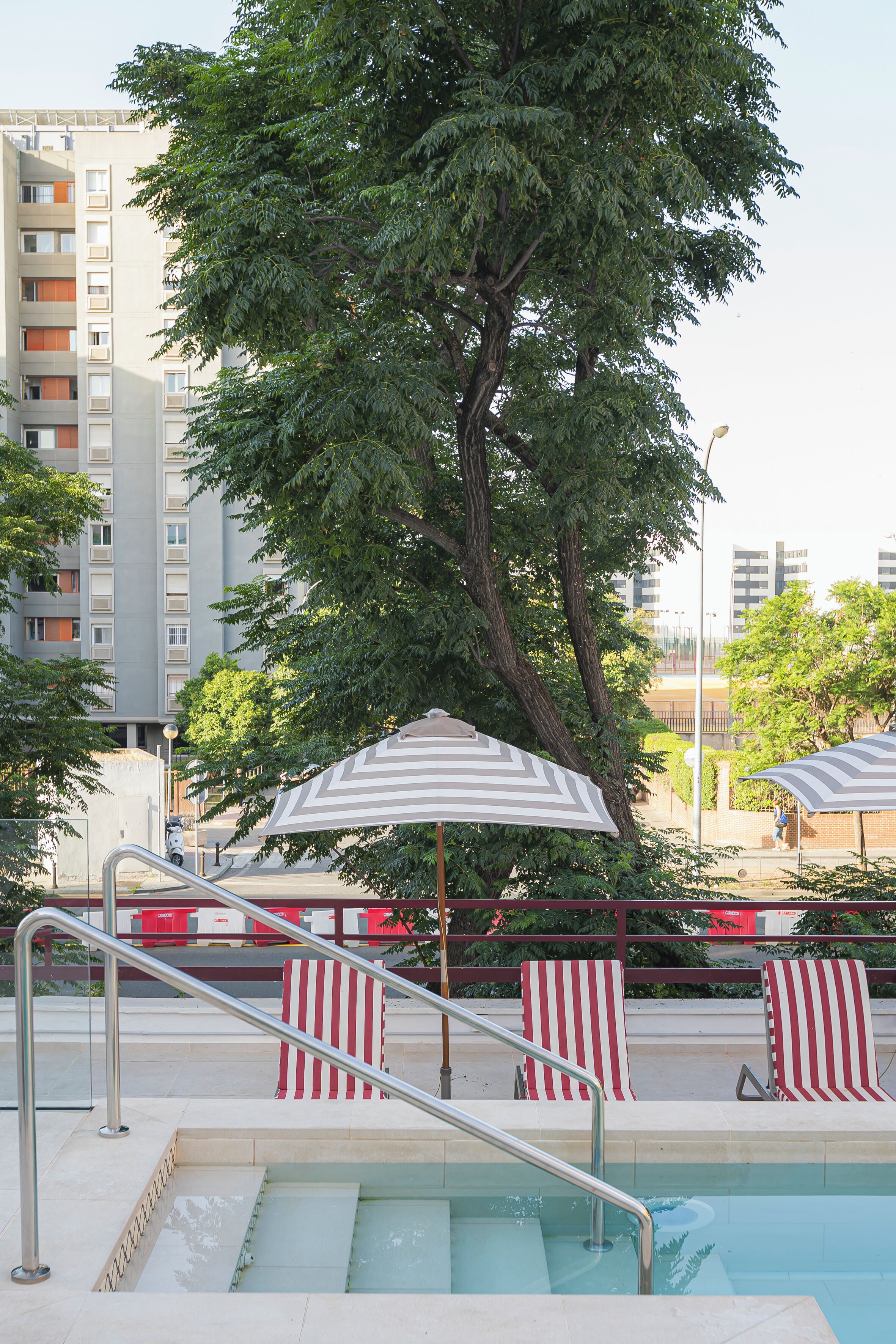 A swimming pool area with striped lounge chairs, a metal staircase, and a large tree in the background.