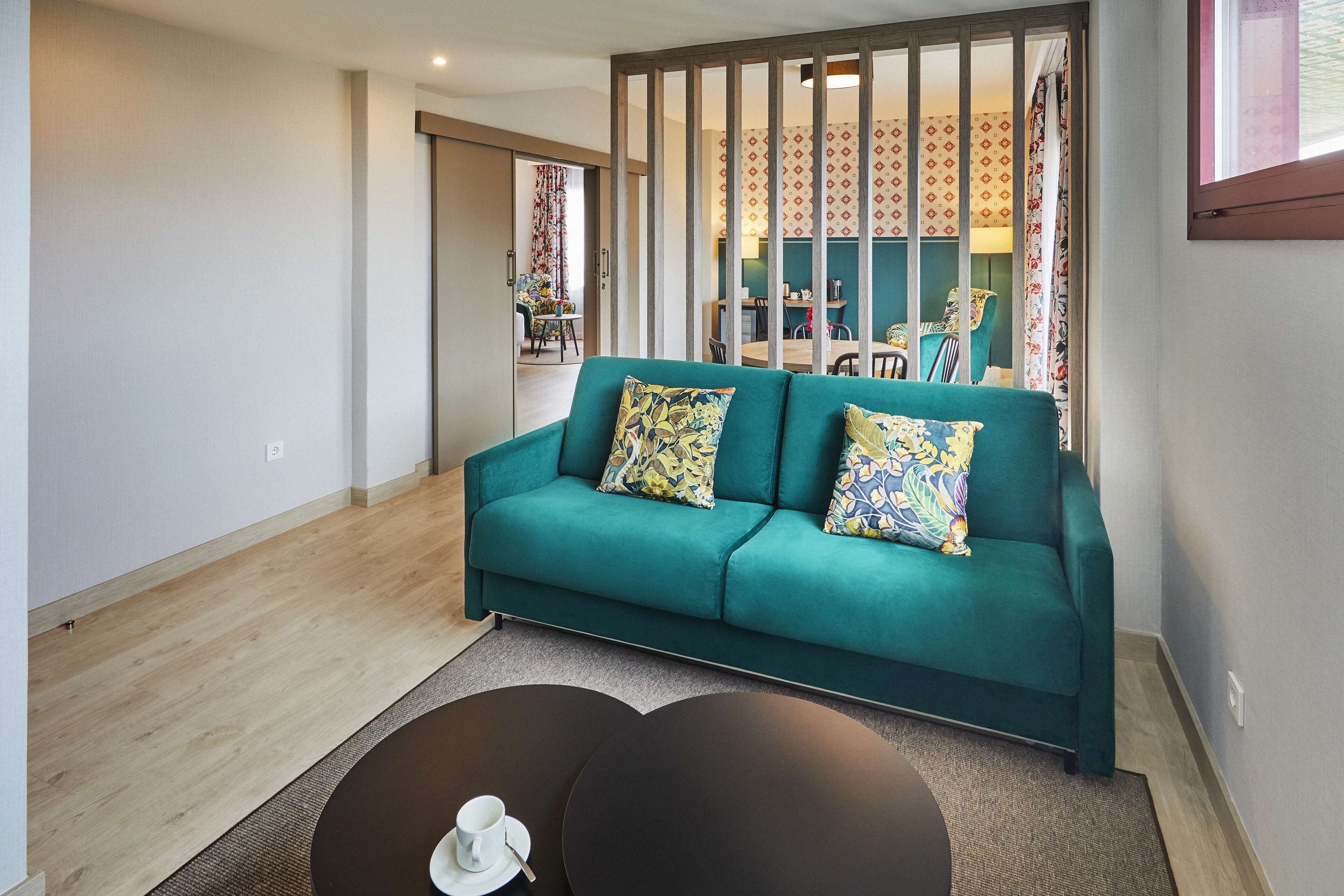 A modern living room with a teal sofa, two patterned cushions, and a coffee table with a cup and saucer.