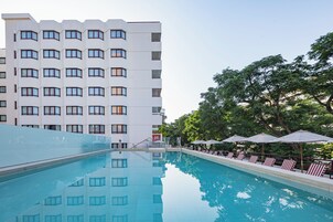 Une piscine entourée d’arbres, avec des chaises longues et des parasols, et un immeuble d’hôtel à plusieurs étages en arrière-plan.