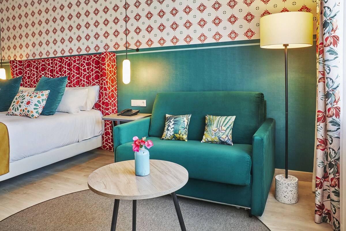 A hotel room with a bed, a green sofa, a small round table with a vase of flowers, and a floor lamp.