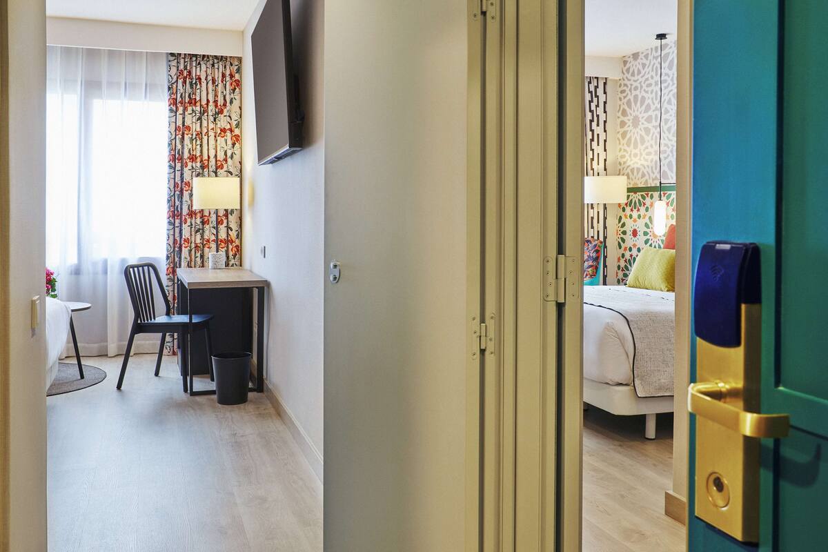 A hotel room with a bed, a desk, a chair, a TV mounted on the wall, and a window with curtains.