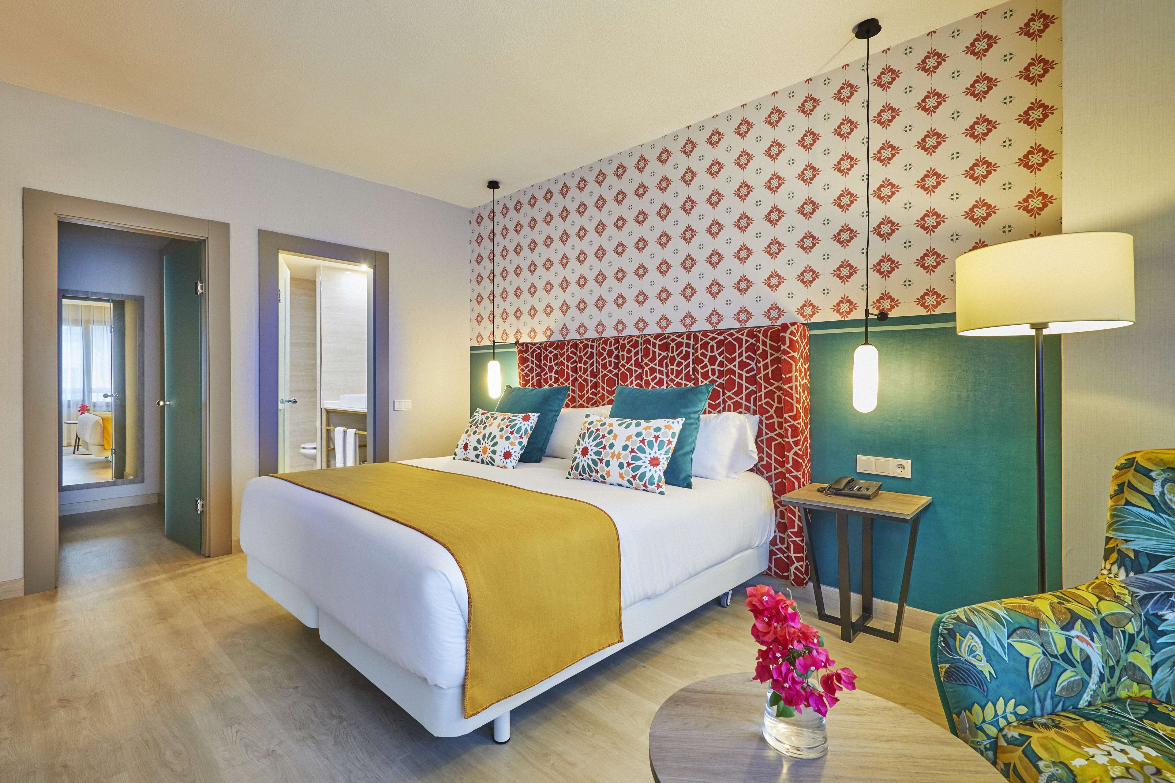 A hotel room with a bed, bedside table, lamp, chair, and a vase with flowers.
