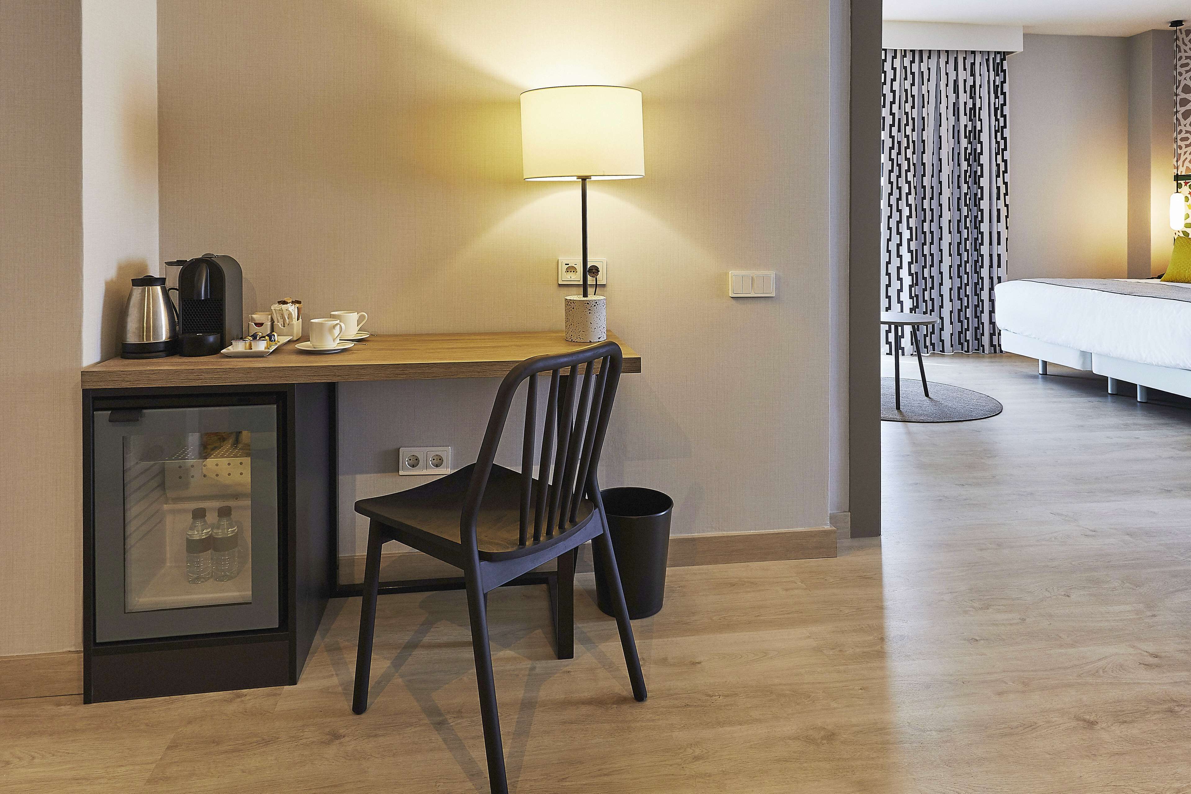 Habitación de hotel con escritorio de madera, una silla, una lámpara, una cama, un refrigerador pequeño y un cubo de basura.