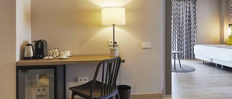 A hotel room with a wooden desk, a chair, a lamp, a bed, a small refrigerator, and a trash bin.