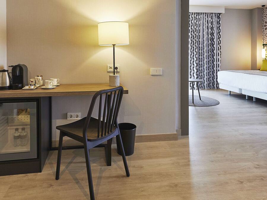Habitación de hotel con escritorio de madera, una silla, una lámpara, una cama, un refrigerador pequeño y un cubo de basura.