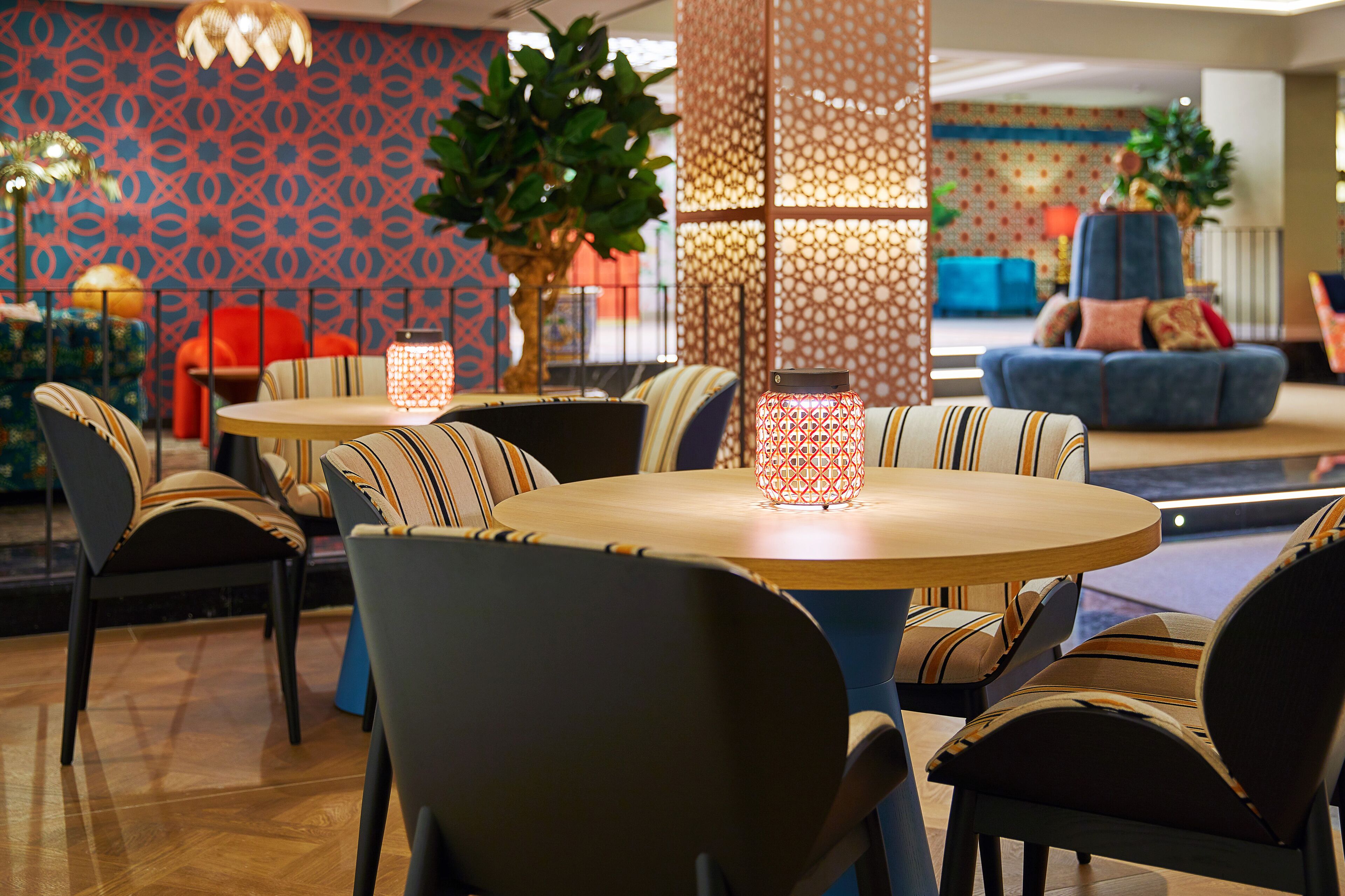 A modern lounge area with round tables and striped chairs, decorative lighting, and patterned wallpaper.