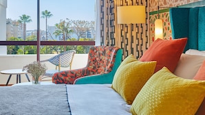 A hotel room with a large window, a bed with colorful pillows, a chair with a floral pattern, and a small table with a vase of flowers. - Hesperia Sevilla (Seville)