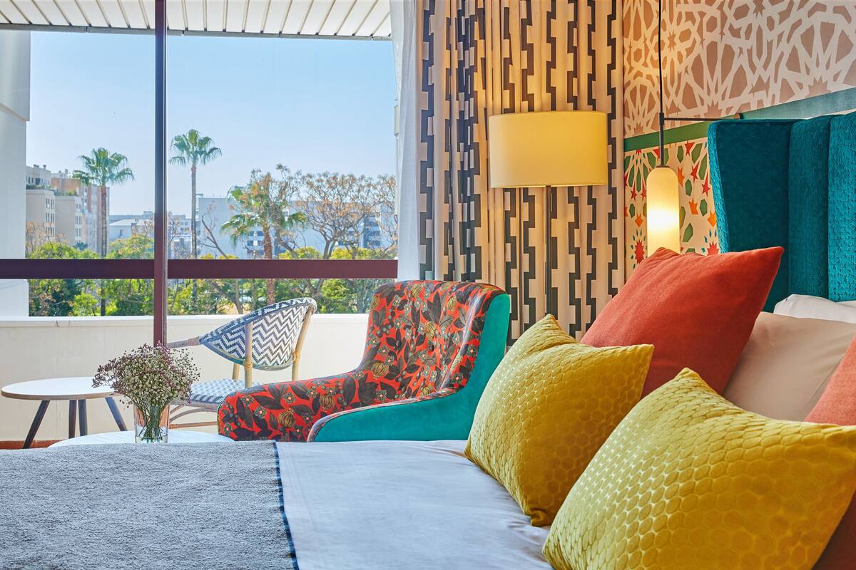 A hotel room with a large window, a bed with colorful pillows, a chair with a floral pattern, and a small table with a vase of flowers.