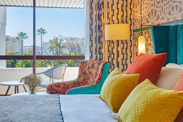 A hotel room with a large window, a bed with colorful pillows, a chair with a floral pattern, and a small table with a vase of flowers. - Hesperia Sevilla (Seville)