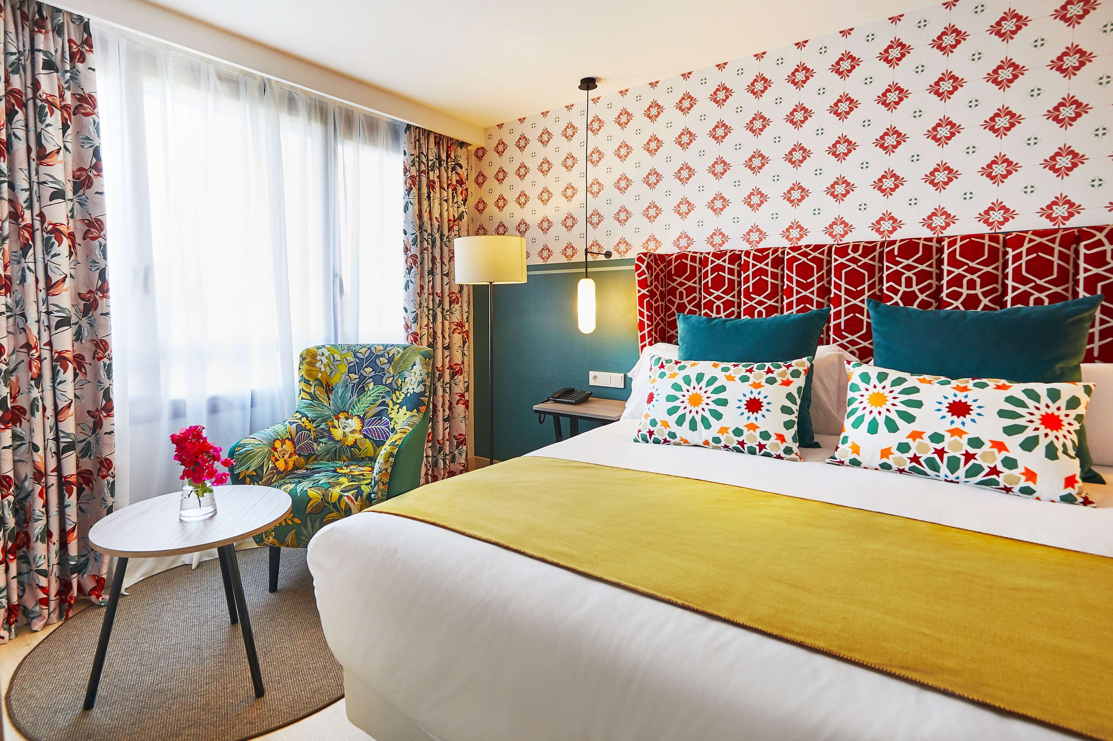 A hotel room with a bed, a chair, a small table with a vase of flowers, and a lamp.