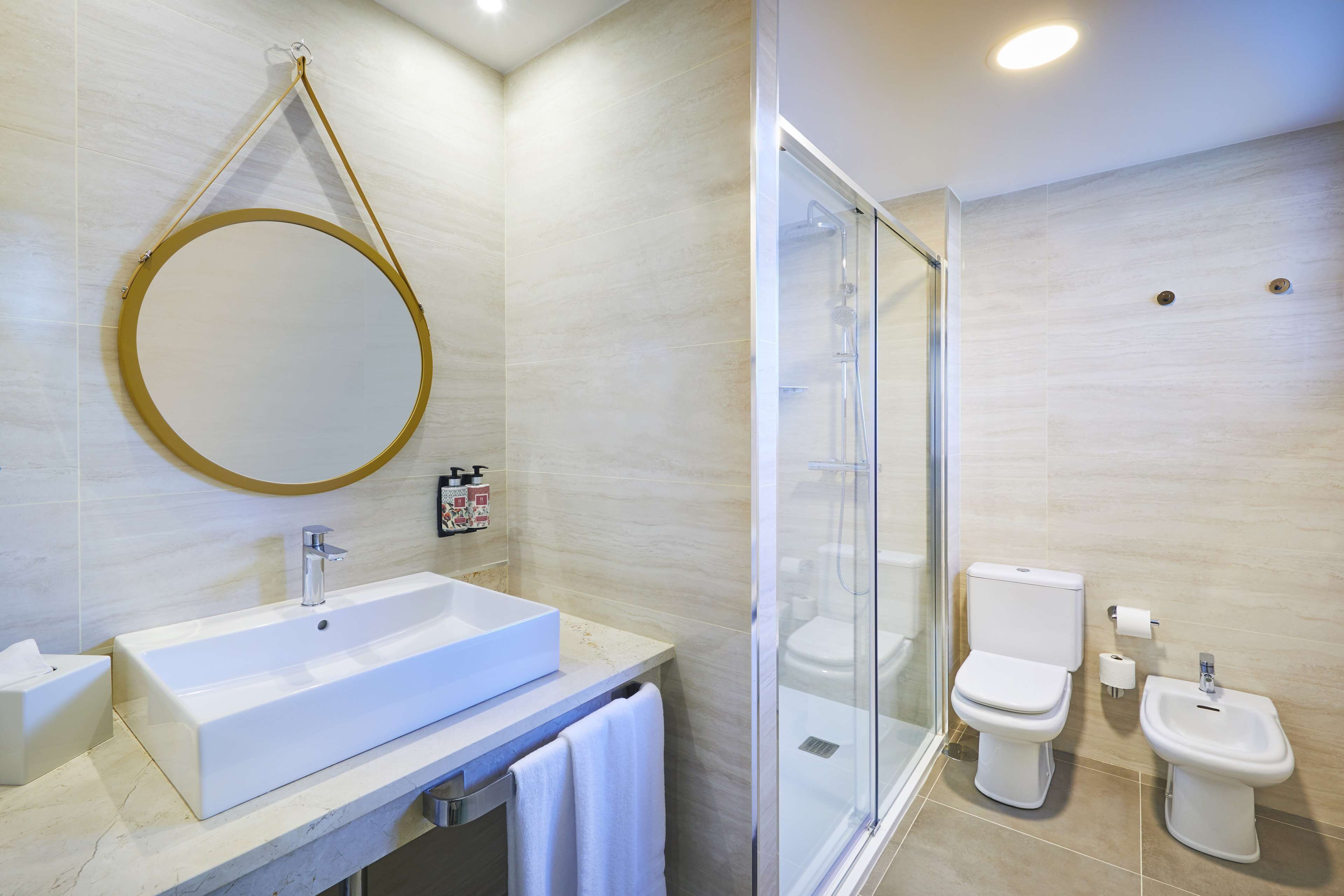 Un baño moderno con un lavamanos grande y rectangular, un espejo redondo, una ducha con mampara de vidrio, un inodoro y un bidé.