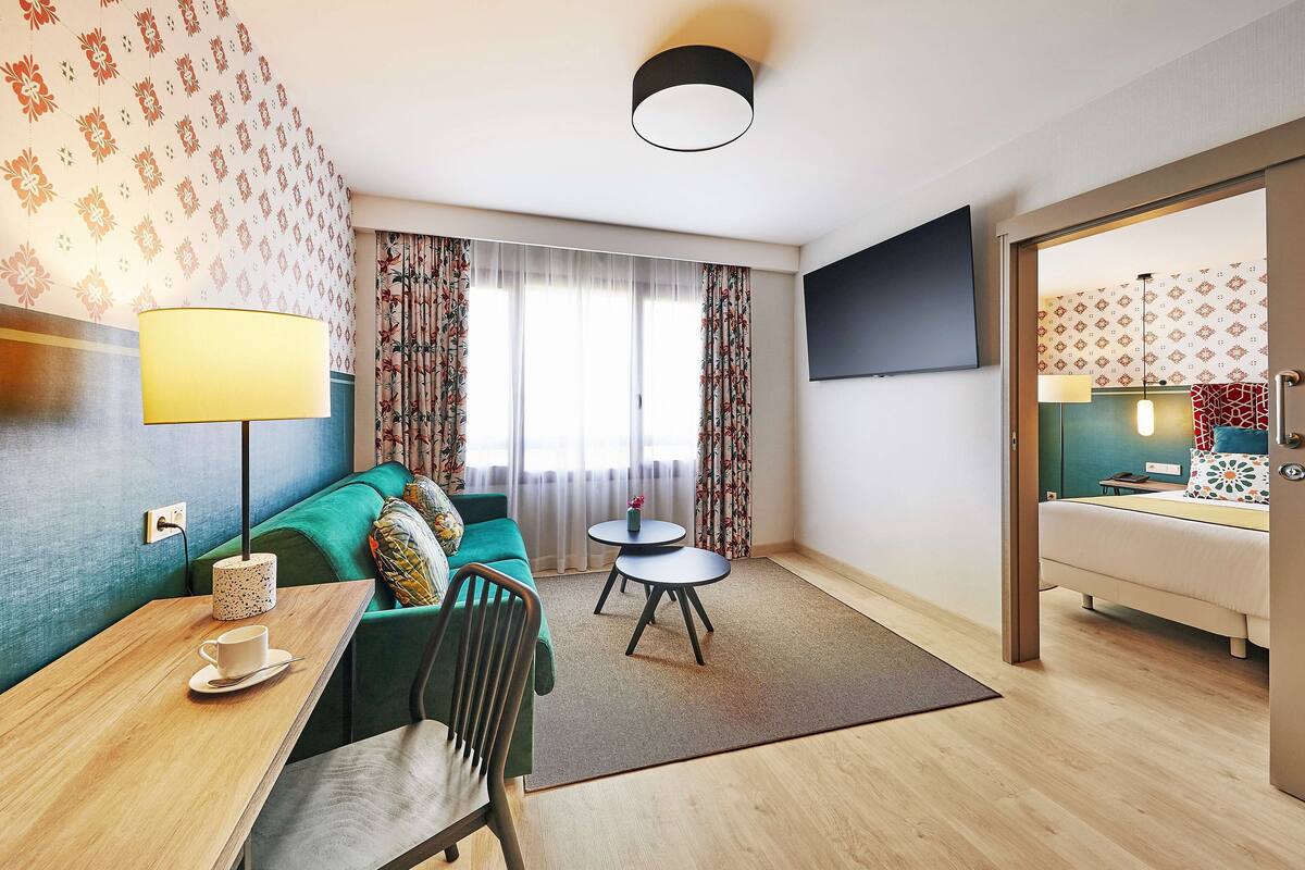 A modern hotel room with a green sofa, a small round table with a cup, a lamp, and a TV mounted on the wall.