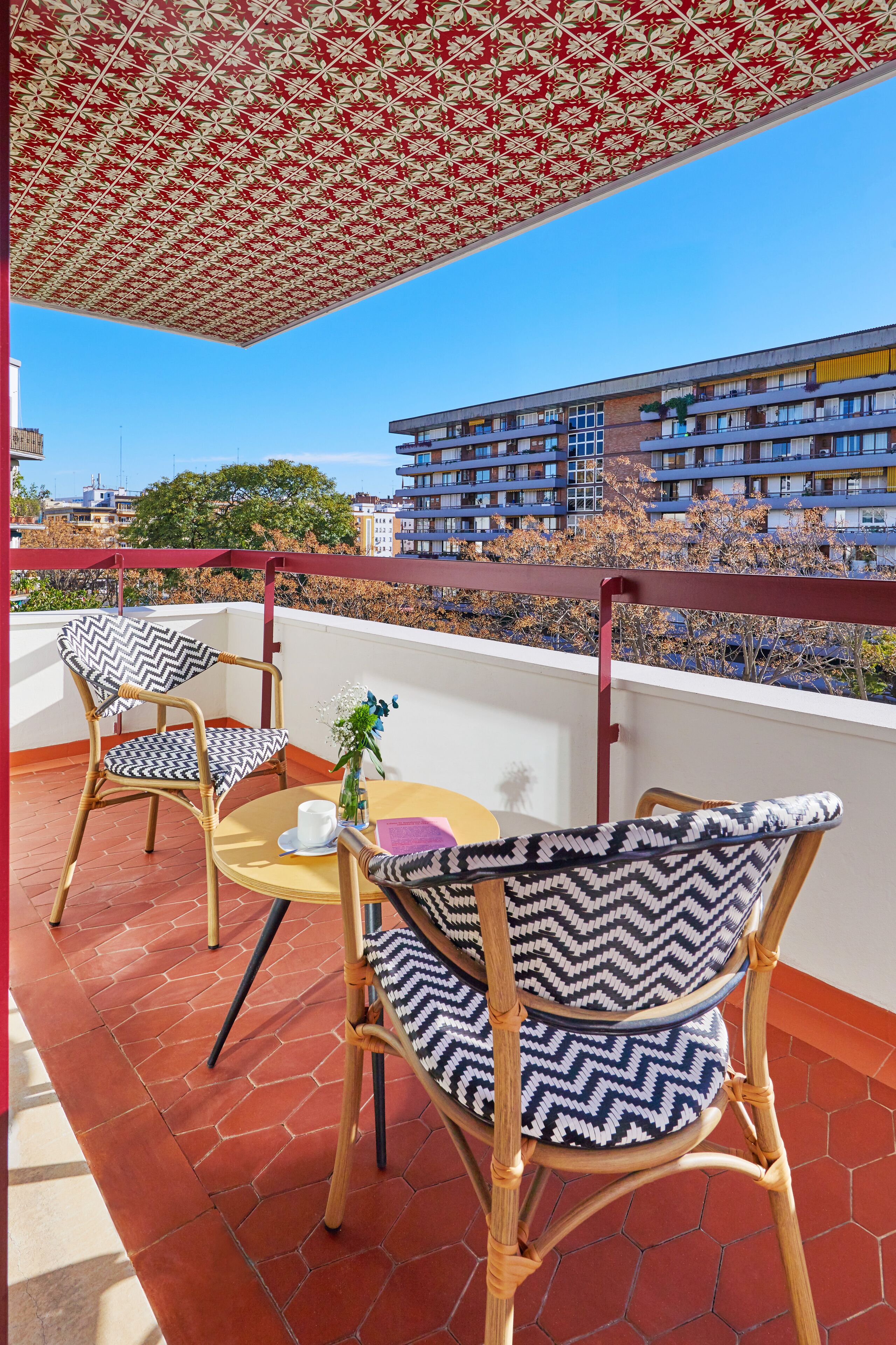 A balcony with wicker chairs and a table, a patterned ceiling, and a view of a multi-story building.