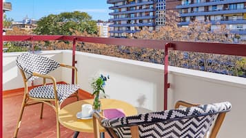 A balcony with wicker chairs and a table, a patterned ceiling, and a view of a multi-story building.