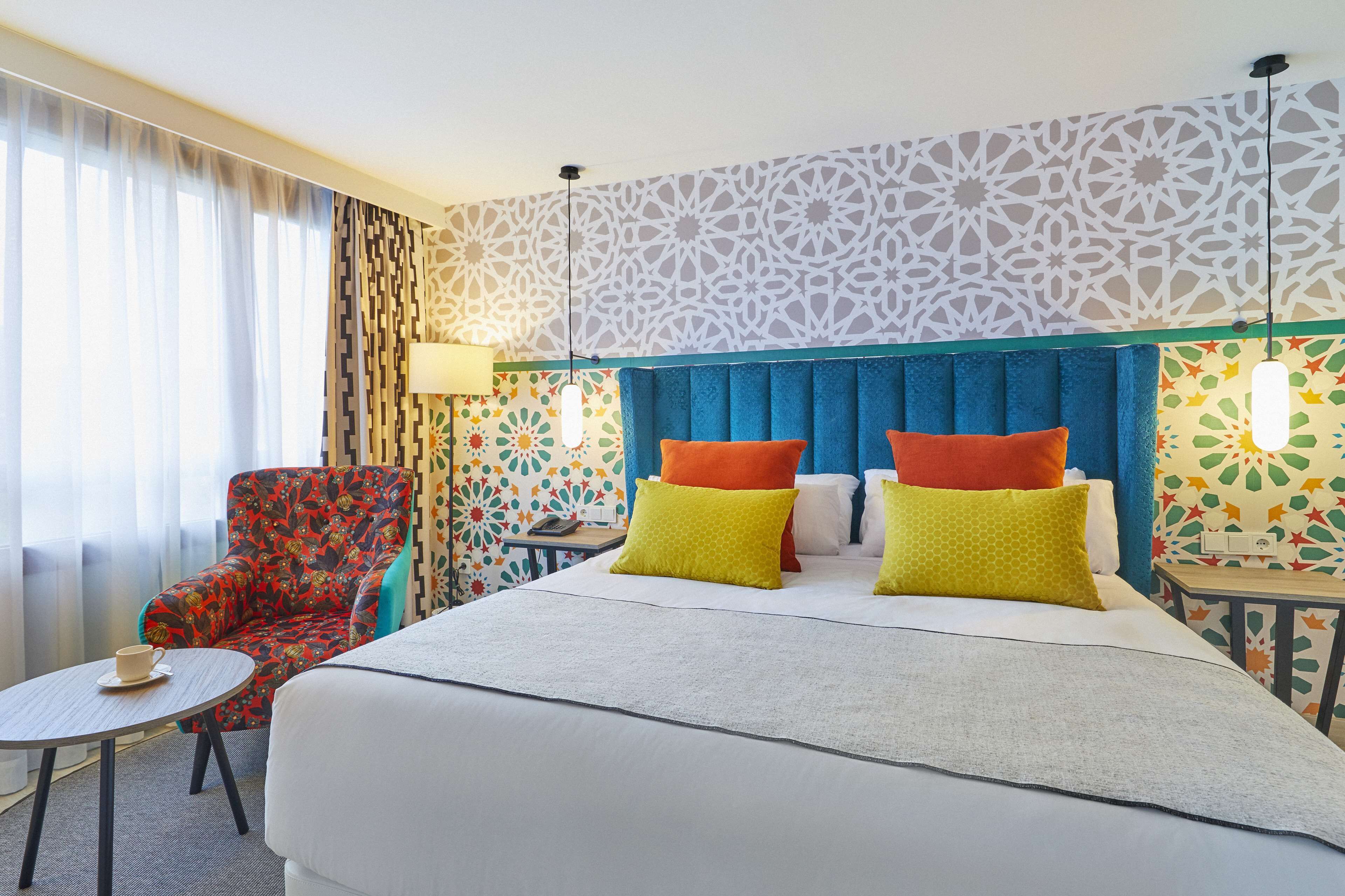 A hotel room with a large bed, a chair, a small table, and a patterned wall.