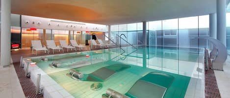 A modern indoor swimming pool with lounge chairs and a glass wall.