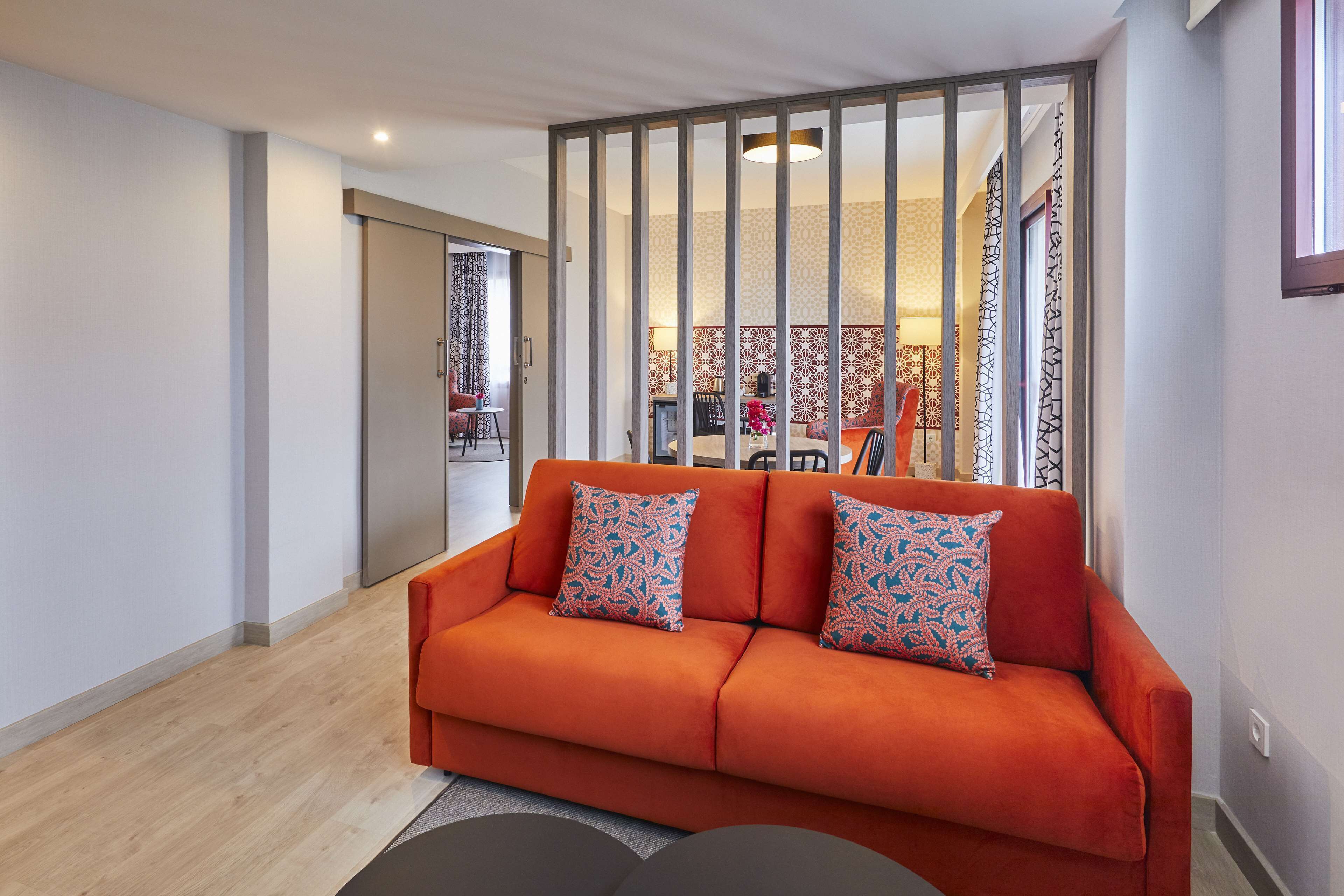 A modern living room with a sofa and patterned cushions, a wooden floor, and a sliding door leading to a dining area.