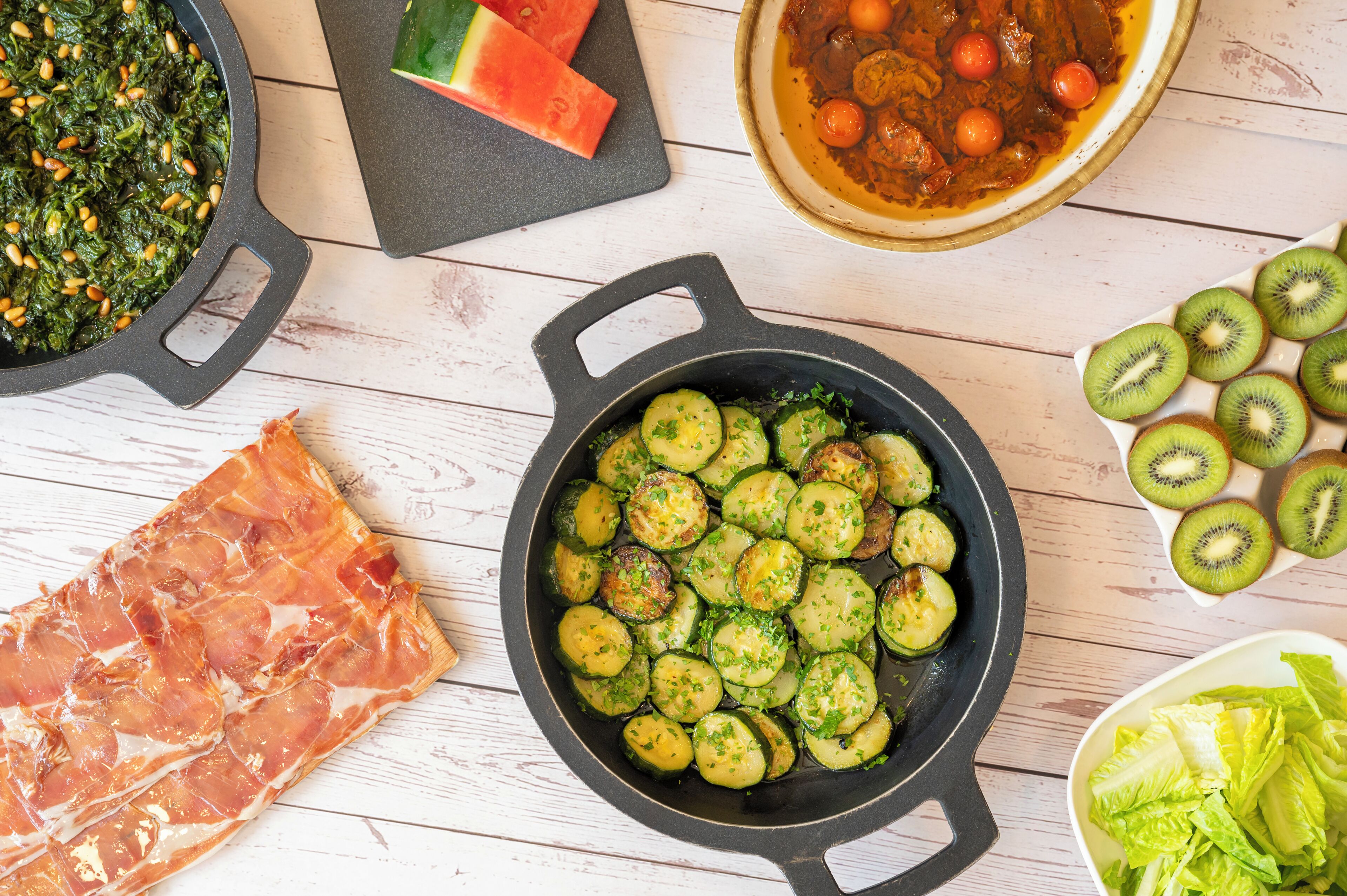 A kitchen with a cast iron skillet, a cutting board with watermelon, a bowl of soup, sliced kiwi, and a plate of prosciutto.