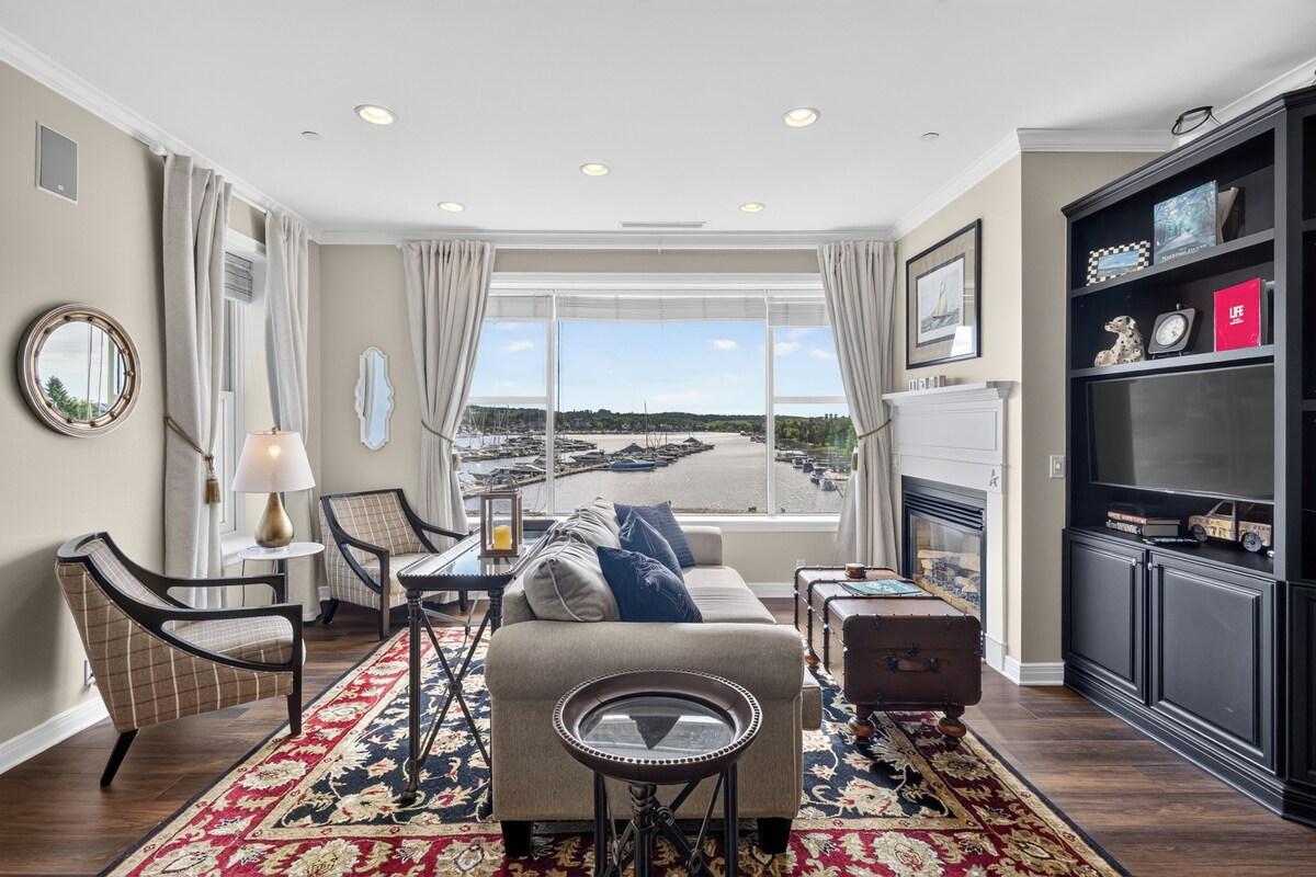 A living room with a view of a marina, a fireplace, and a bookshelf with various items.