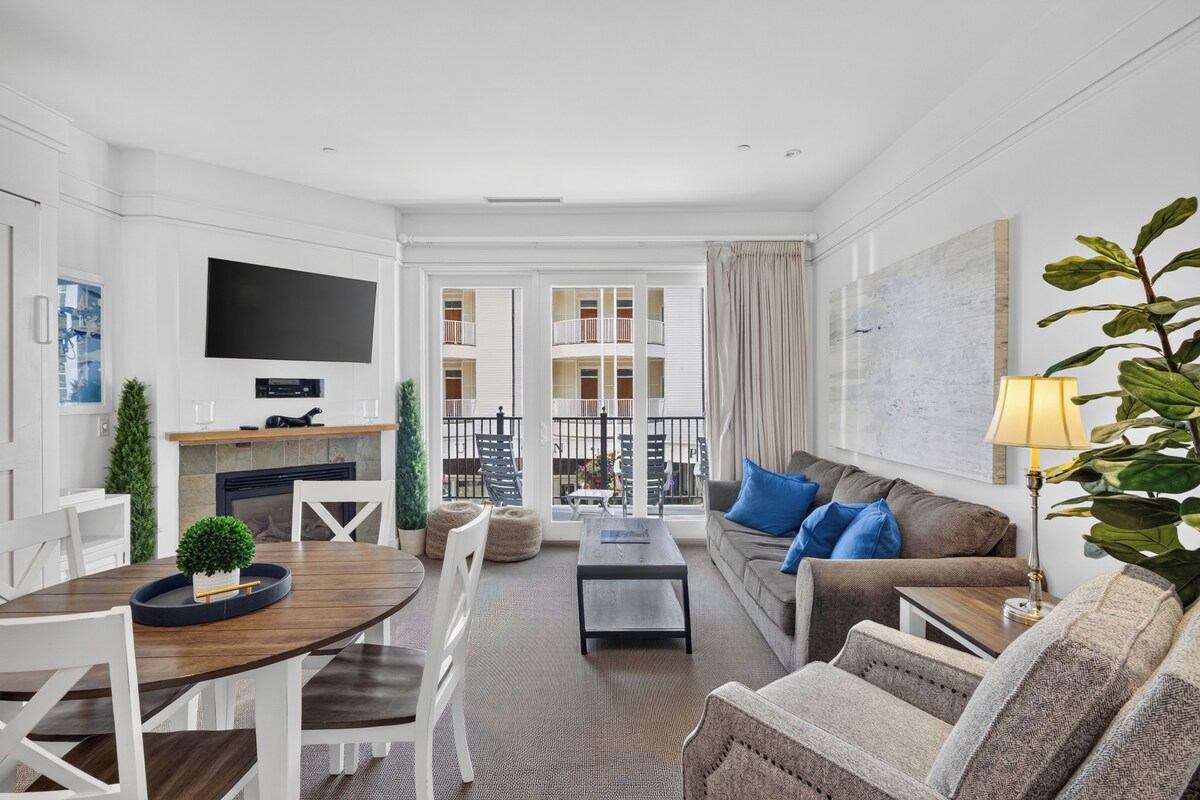 A modern living room with a flat-screen TV, a fireplace, a dining area with a wooden table and chairs, and a balcony with a view.