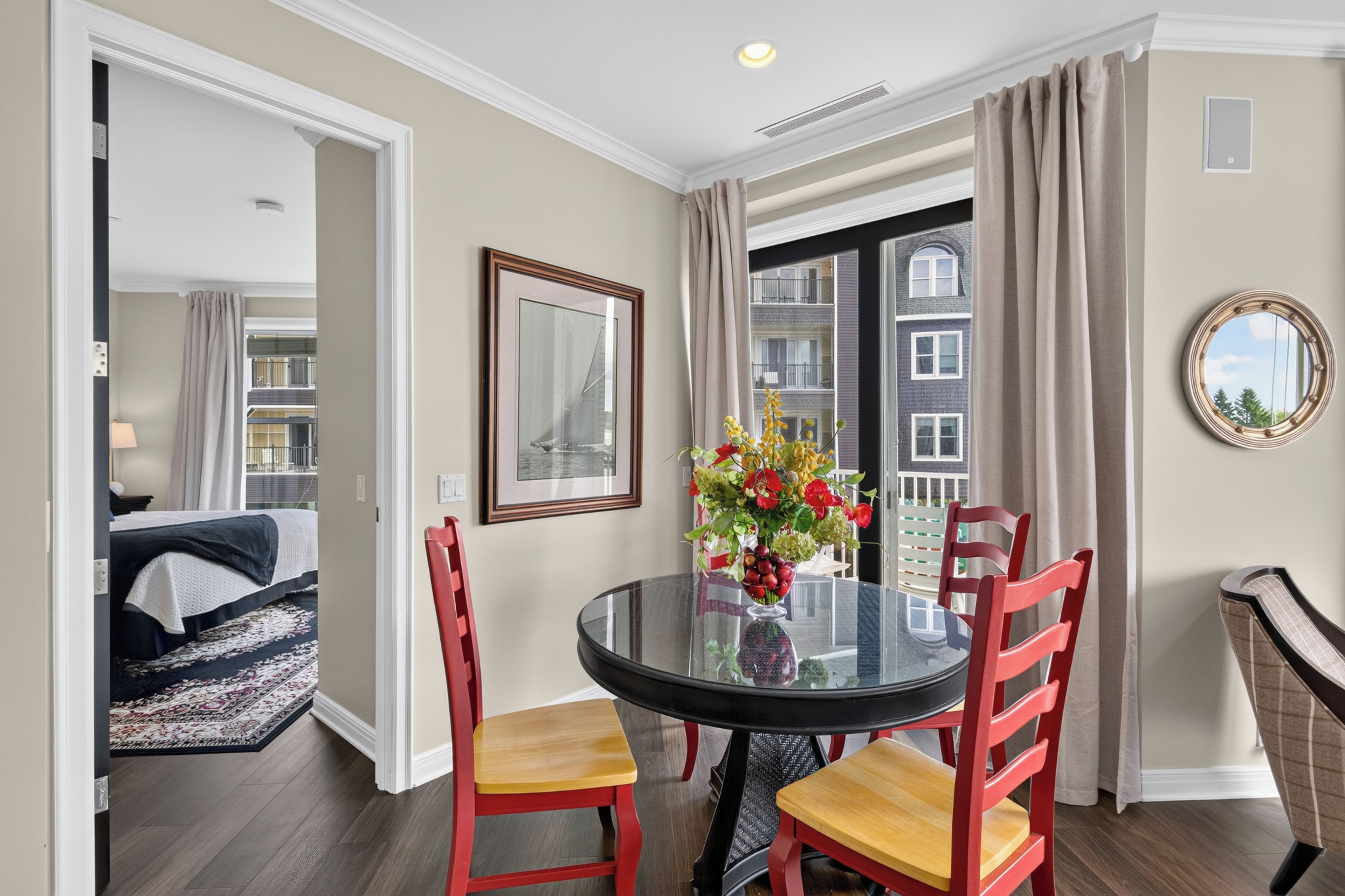 A dining area with a glass table, red chairs, a vase of flowers, a framed picture, and a view of a building through a window.