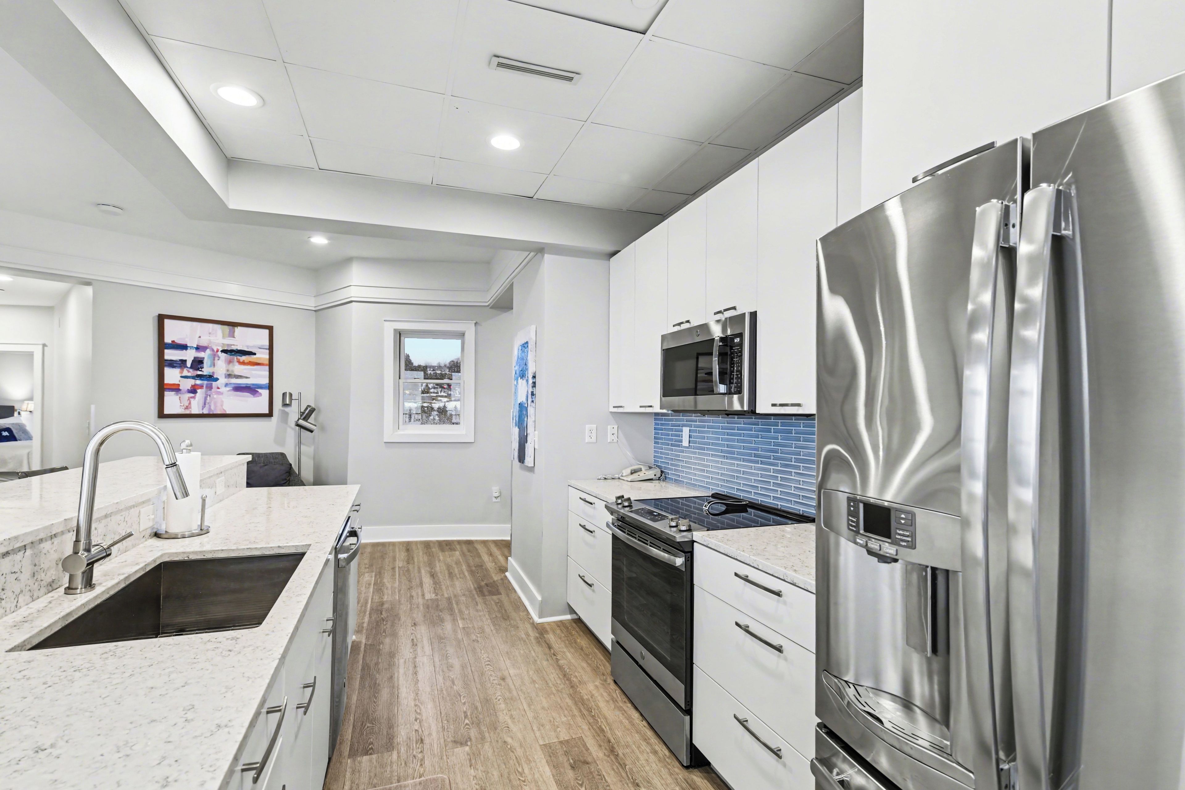 A modern kitchen with stainless steel appliances, a marble countertop, and a view to the outside.
