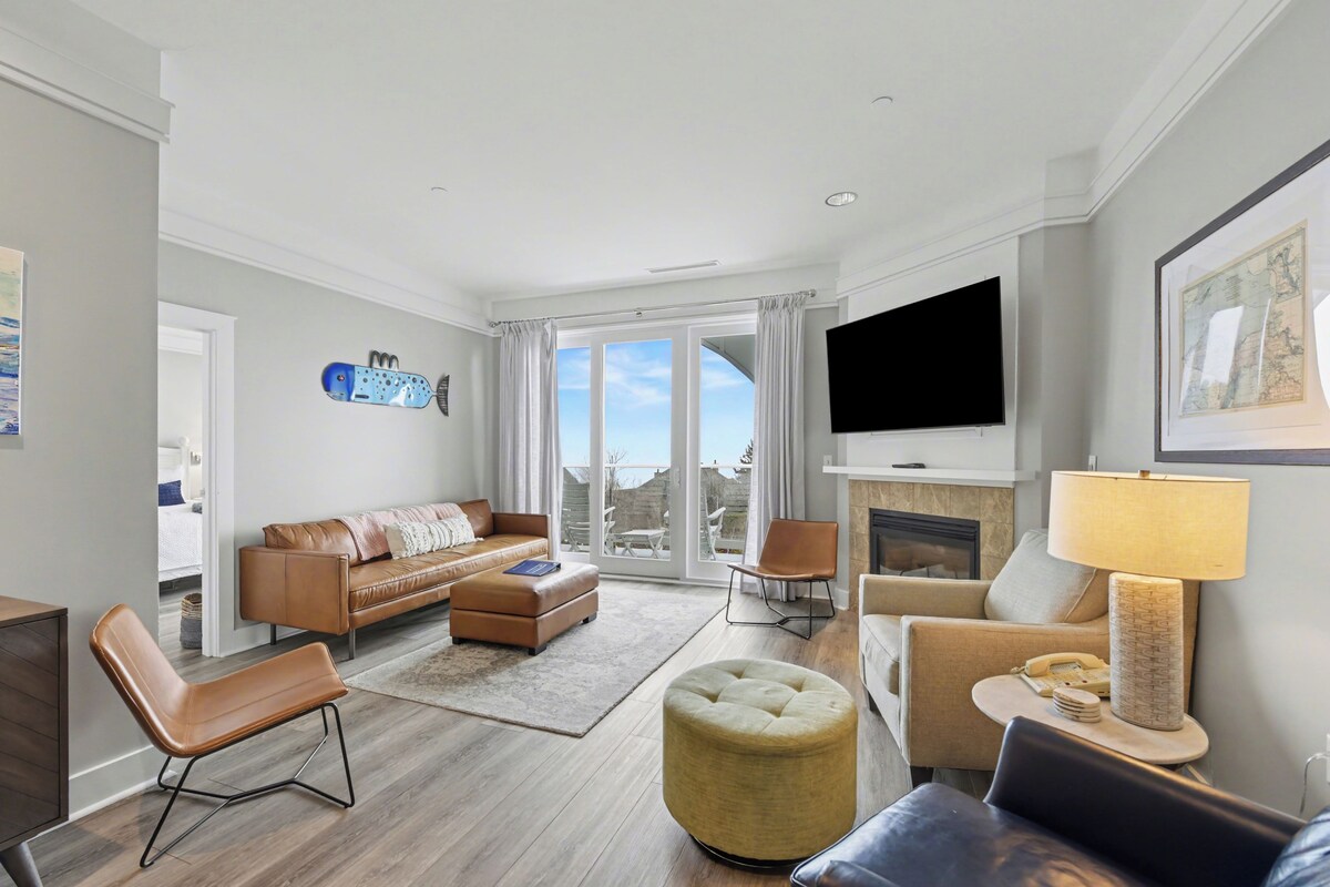 A modern living room with a sofa, ottoman, chairs, a flat-screen TV, and a fireplace.