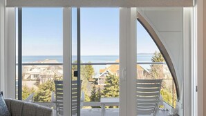 A room with a large sliding glass door leading to a balcony with a view of the sea and houses. - Village Suites Bay Harbor (Petoskey)