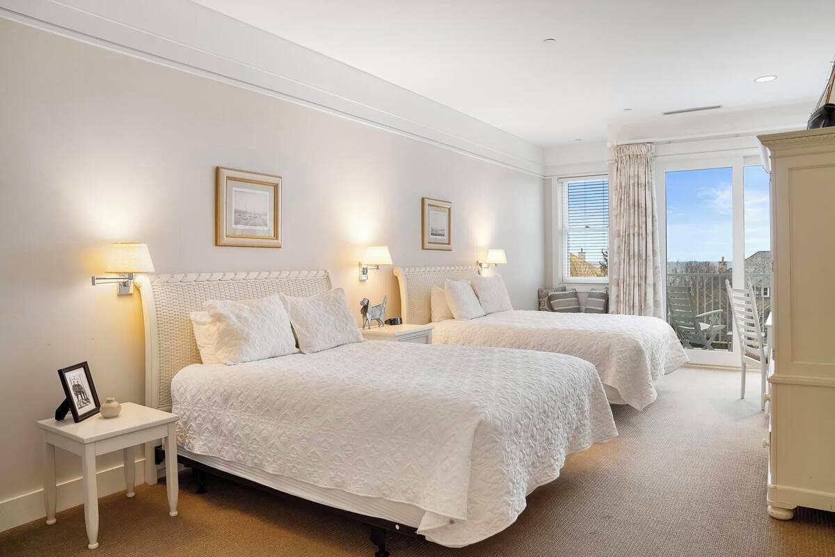 A hotel room with two beds, a small table, bedside lamps, a framed picture, and a view of the city through large windows.