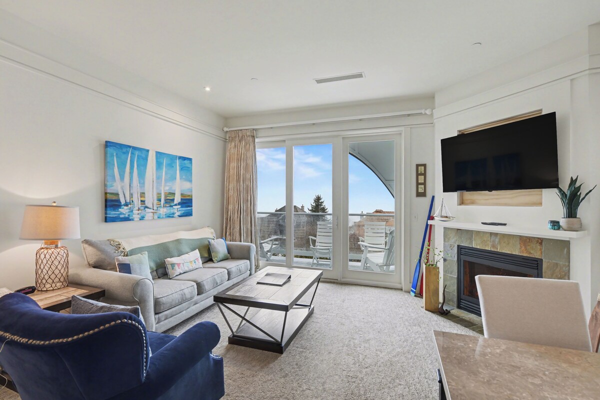 A living room with a sofa, coffee table, TV, fireplace, and large windows.