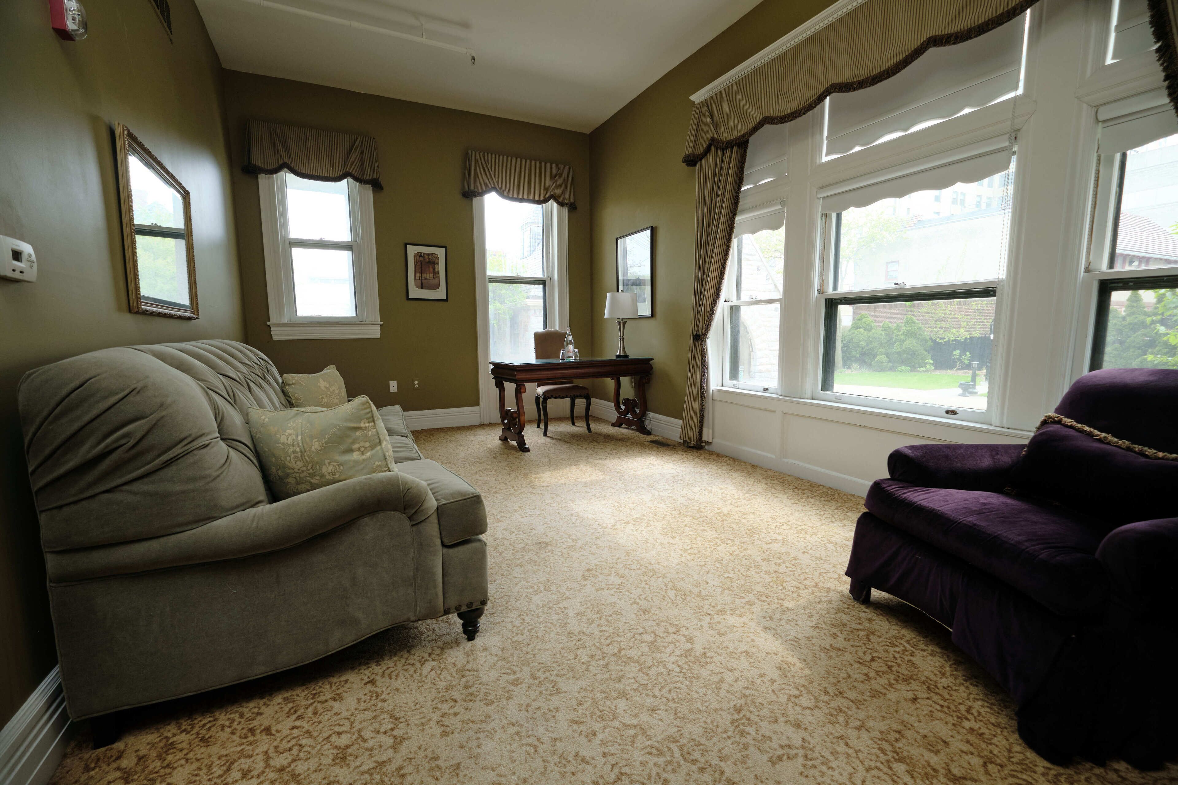 A room with a beige carpet, a beige sofa, a purple armchair, a small wooden table with a lamp, and two windows with curtains.