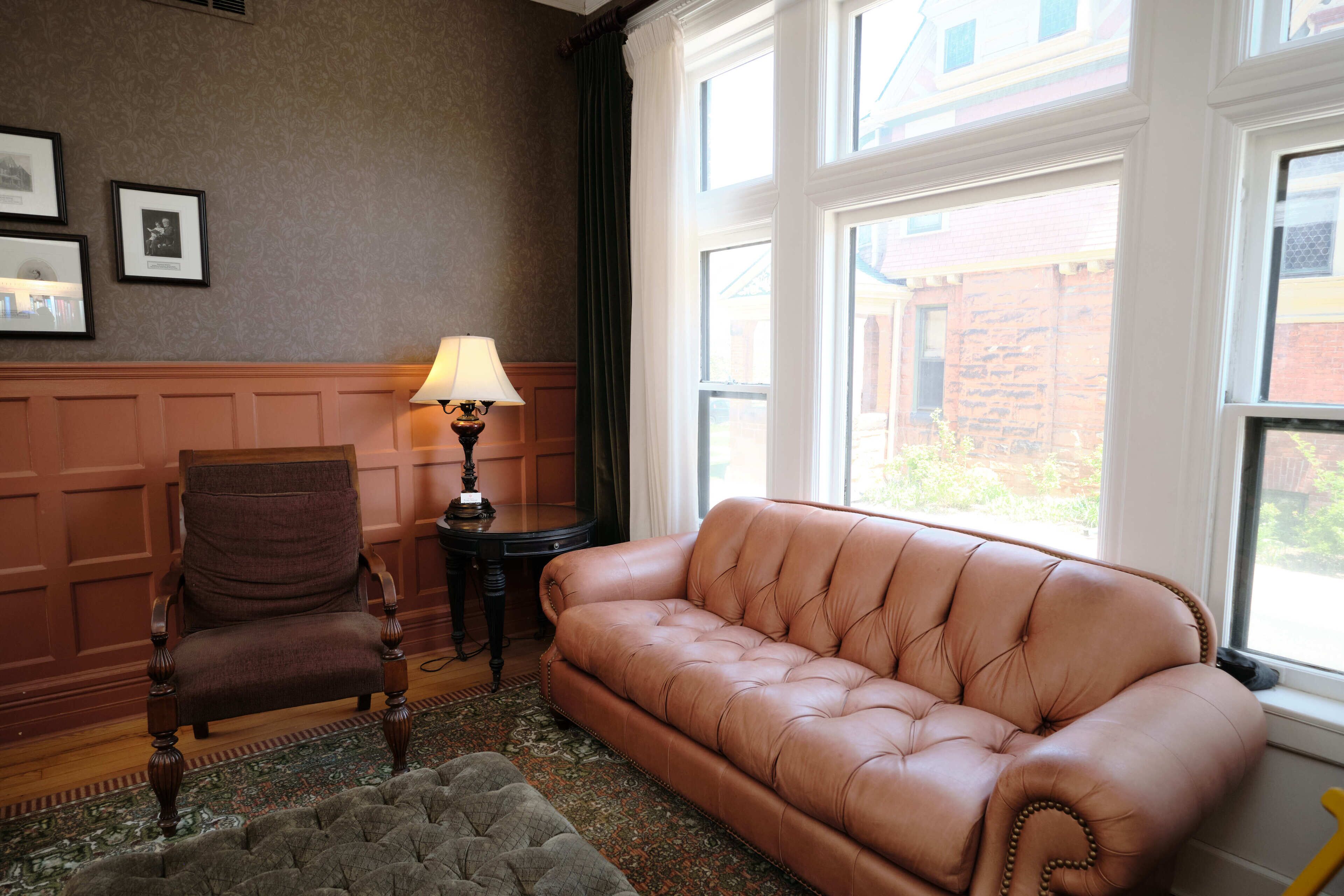 A room with a brown leather sofa, a wooden chair, a small table with a lamp, and a patterned rug.